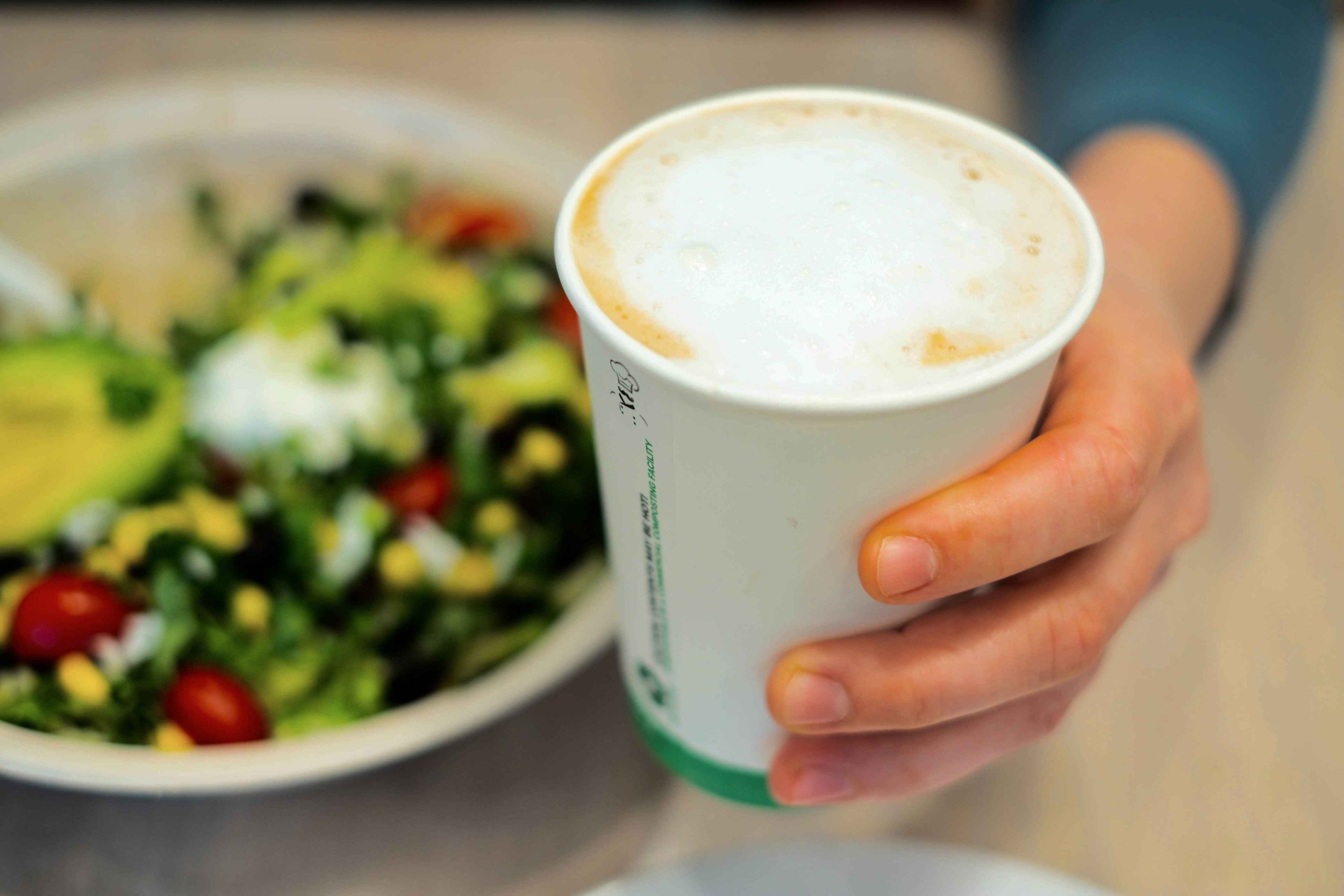 A person holding a white paper cup of coffee or cappuccino. There is a bowl of salad with greens, cherry tomatoes, and possibly avocado in the background.