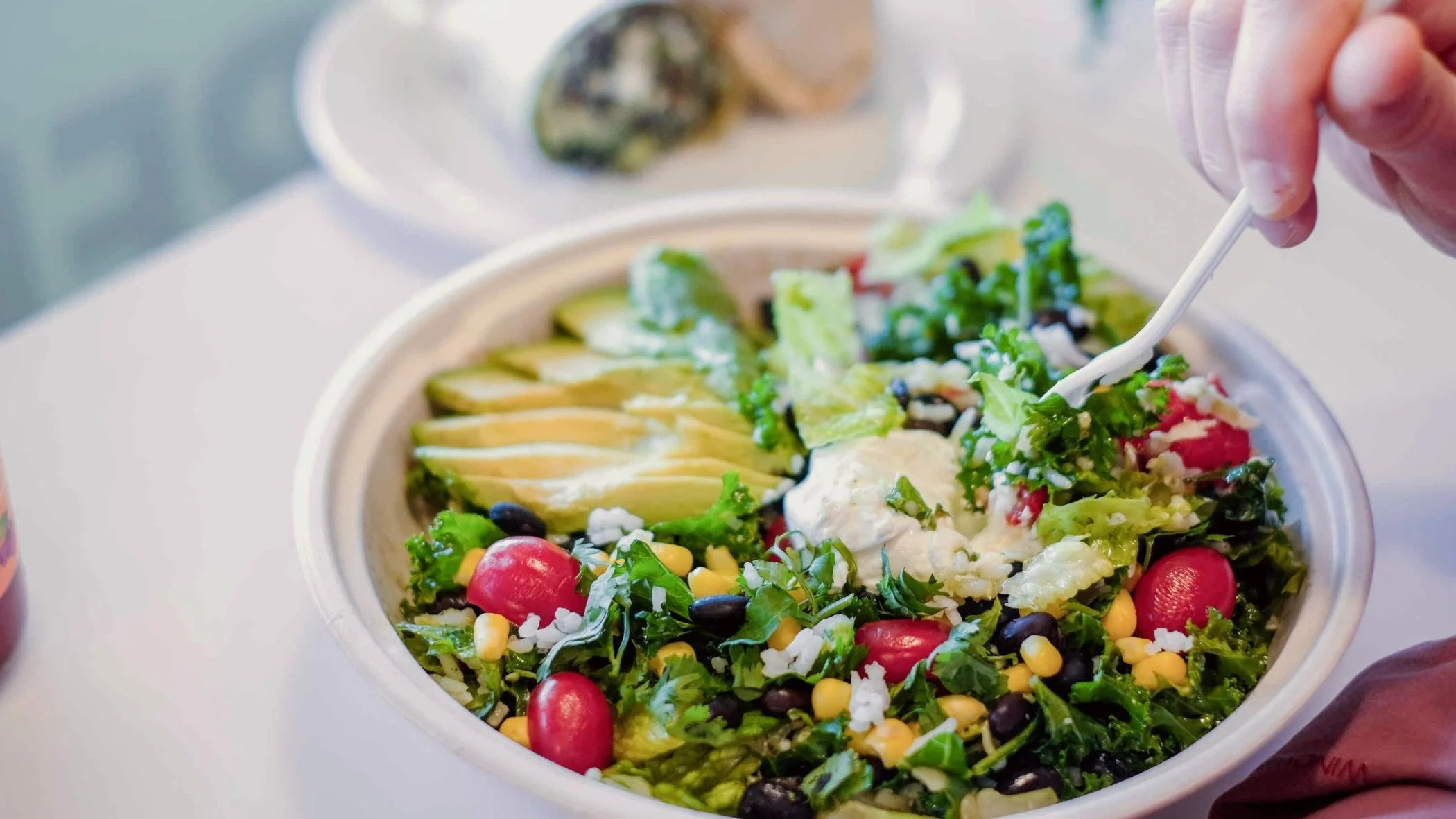 A bowl of mixed salad with cherry tomatoes, corn, lettuce, avocado slices, black beans, shredded cheese, and ranch dressing, with a person using a fork to pick up some salad.