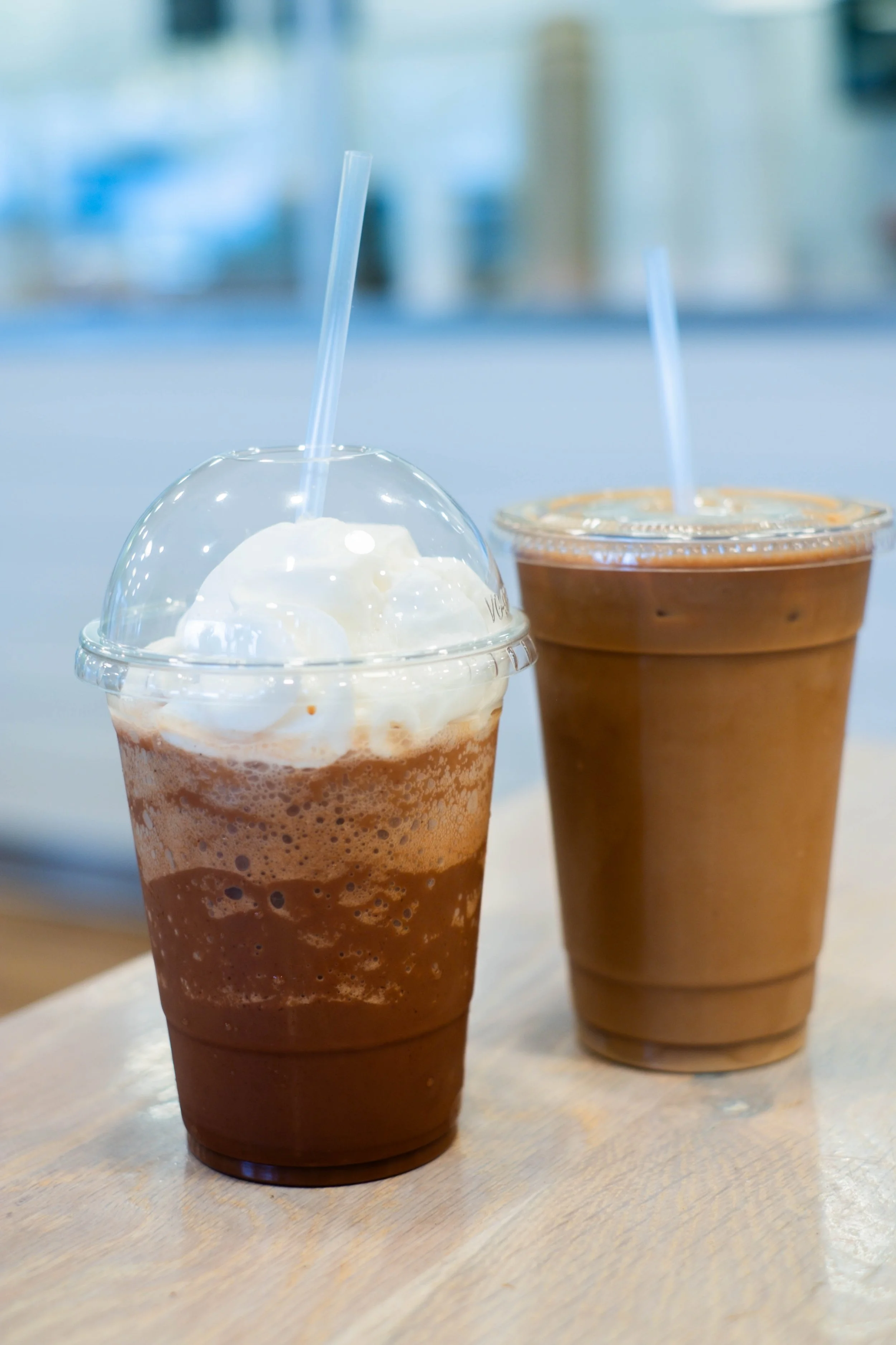 Two iced coffee drinks in clear plastic cups with dome lids and straws, placed on a light wooden table.