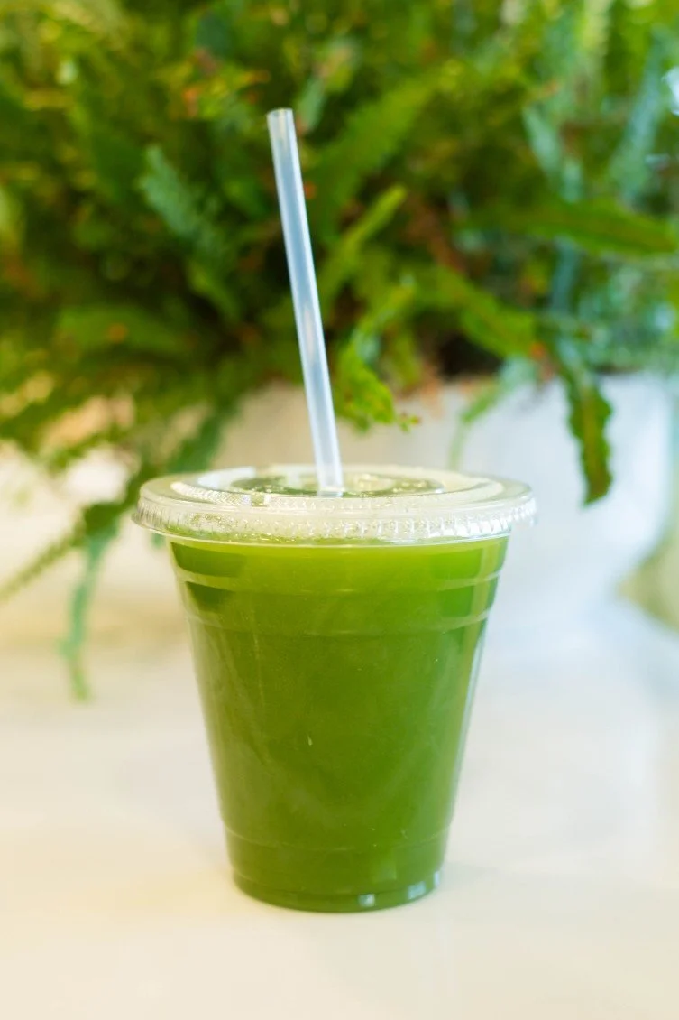 A clear plastic cup filled with green juice, with a clear straw and a plastic lid, placed on a white surface with green plants in the background.