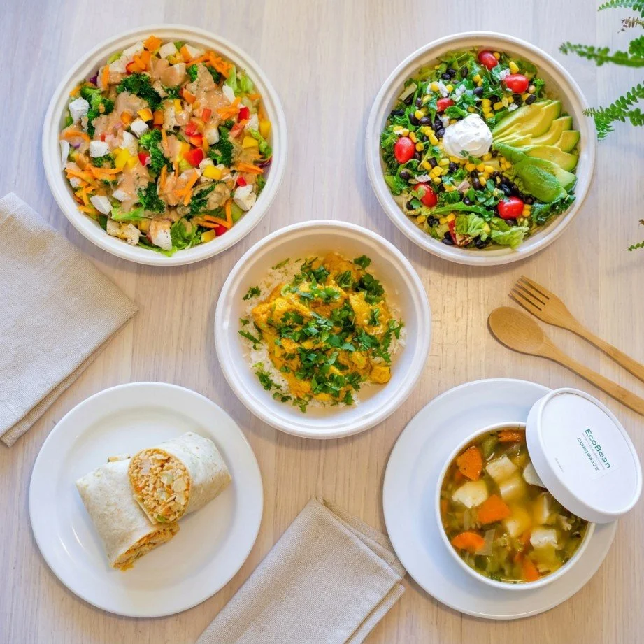 A meal with five dishes including a vegetable salad, another mixed greens salad with cherry tomatoes and avocado, a small bowl of scrambled eggs garnished with herbs, a wrapped breakfast burrito, and a bowl of vegetable soup.