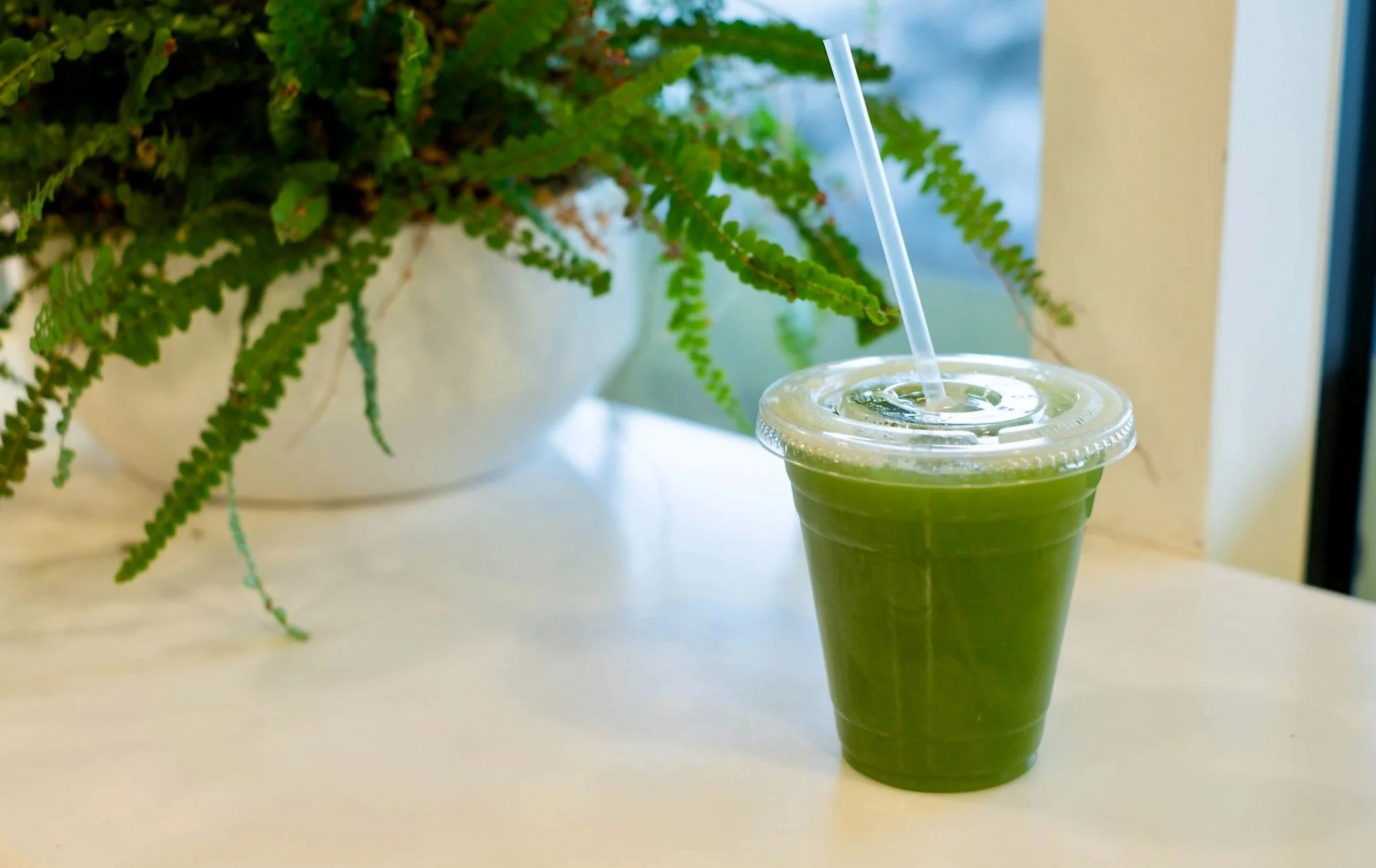 A green juice in a clear plastic cup with a lid and straw on a white table, with a large potted fern plant in the background.