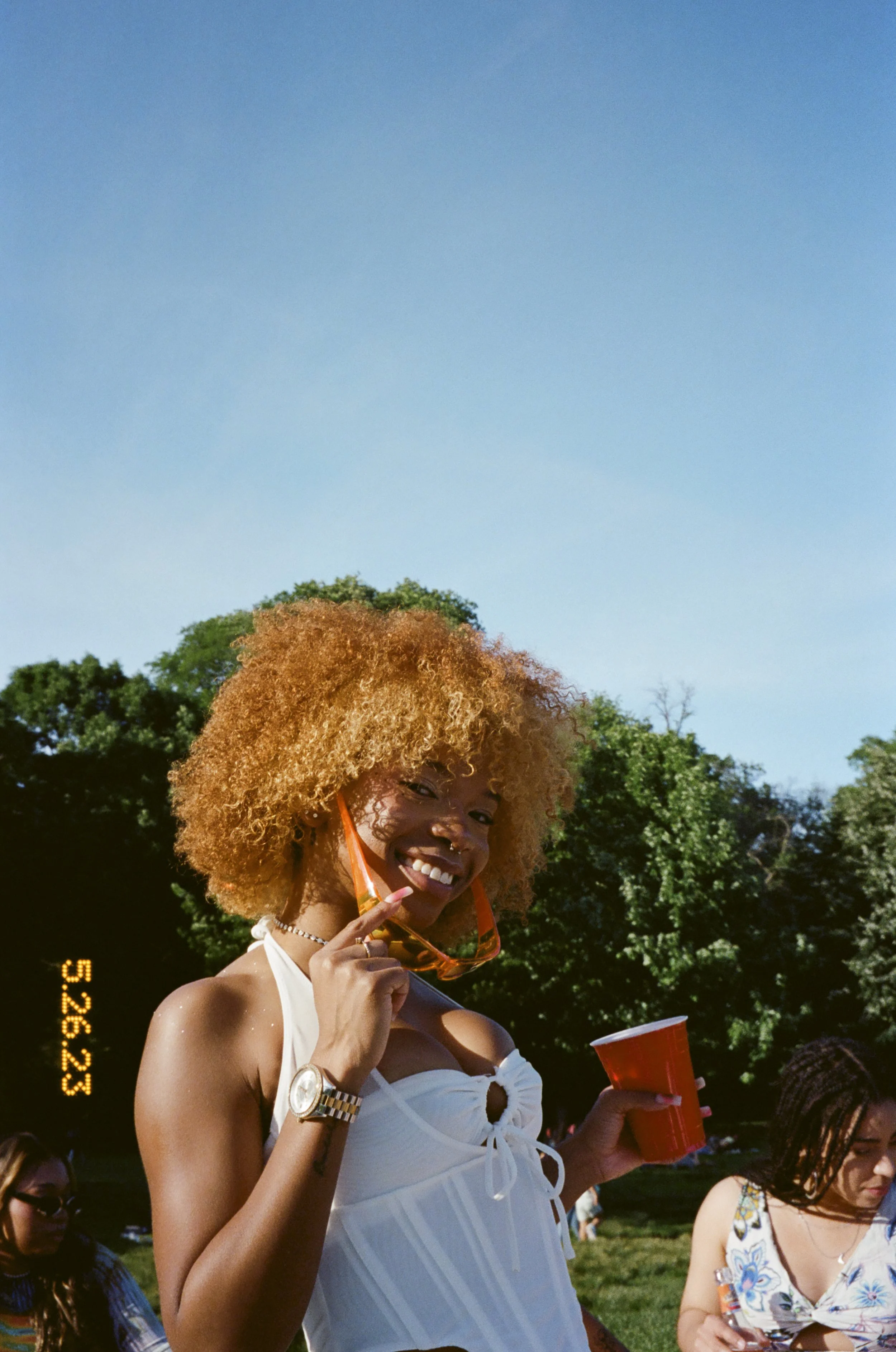 A young woman with curly blonde hair smiling and holding a red cup outdoors on a sunny day, wearing a white top and glasses, with other people and green trees in the background.