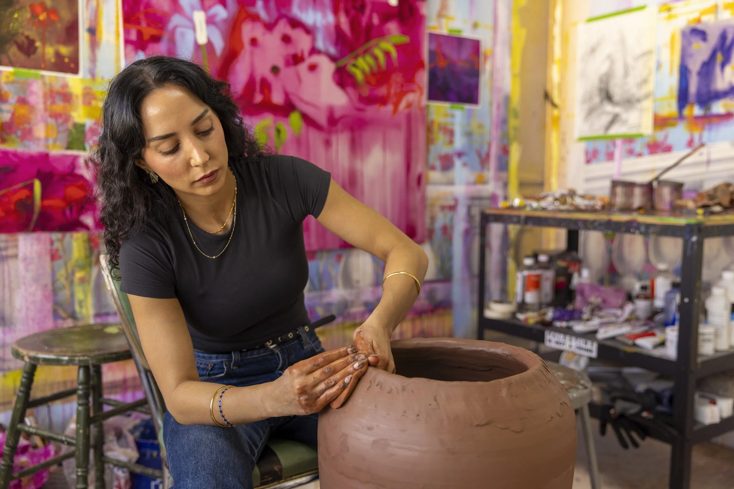 A woman with curly dark hair, wearing a black t-shirt and jeans, shaping clay on a pottery wheel in a colorful art studio.
