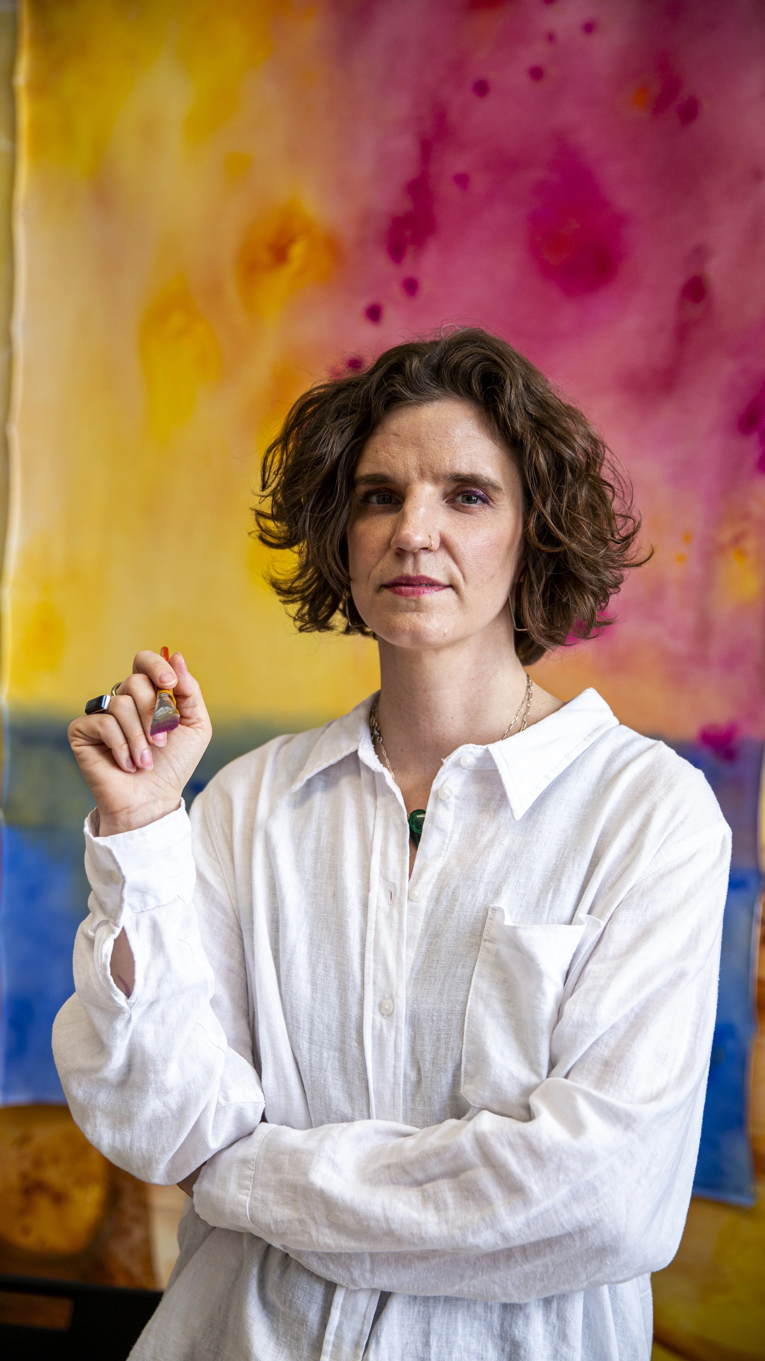 A woman with short, curly brown hair, wearing a white button-up shirt, stands in front of a colorful abstract background. She holds a paintbrush in her right hand, with her arms crossed, looking directly at the camera.