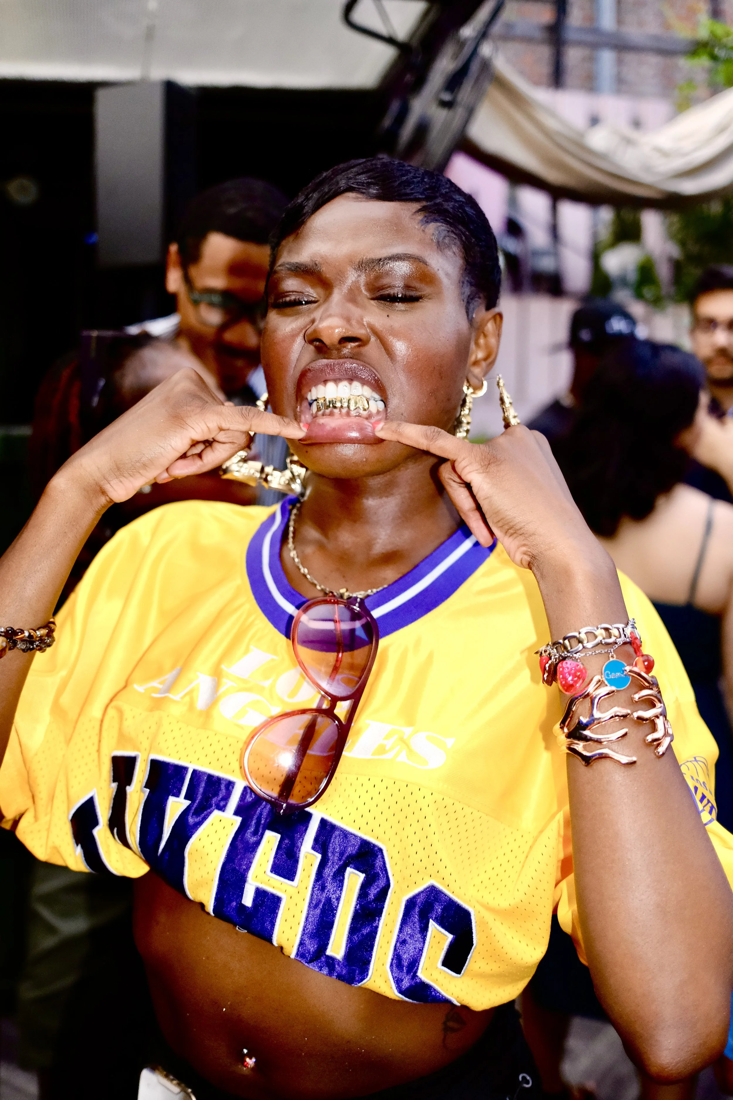 Woman with short hair showing gold grillz on her teeth, wearing a yellow sports jersey and sunglasses hanging from her collar, adjusting her grillz with her fingers, wearing various jewelry, in a lively outdoor setting.