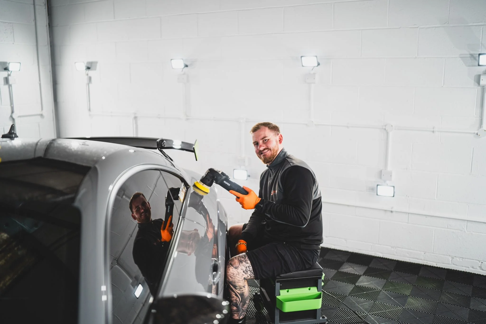 A man with tattoos on his leg, wearing gloves, is holding a polishing tool and working on a silver car in a well-lit garage with white walls and black-and-white checkered flooring.