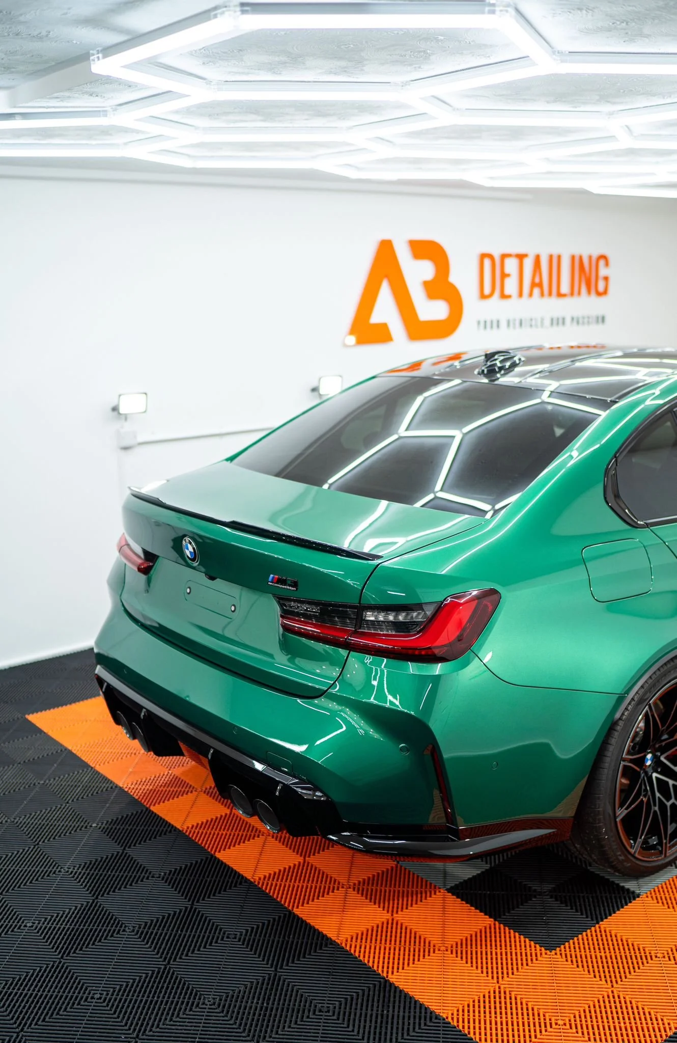 Green BMW M4 sports car in detailing garage with white walls, illuminated ceiling, and checkered floor with orange accents.