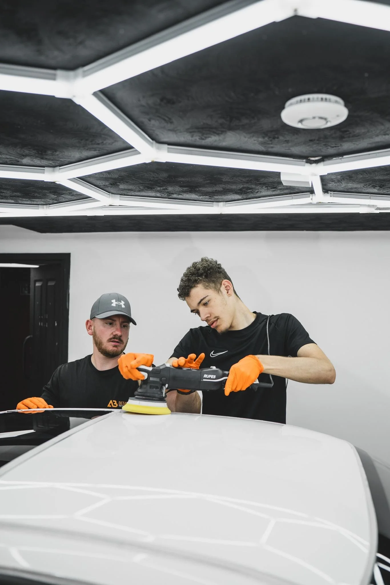 Two men wearing black shirts and orange gloves working together on a car at a detailing workshop, with a man in a gray Under Armour cap