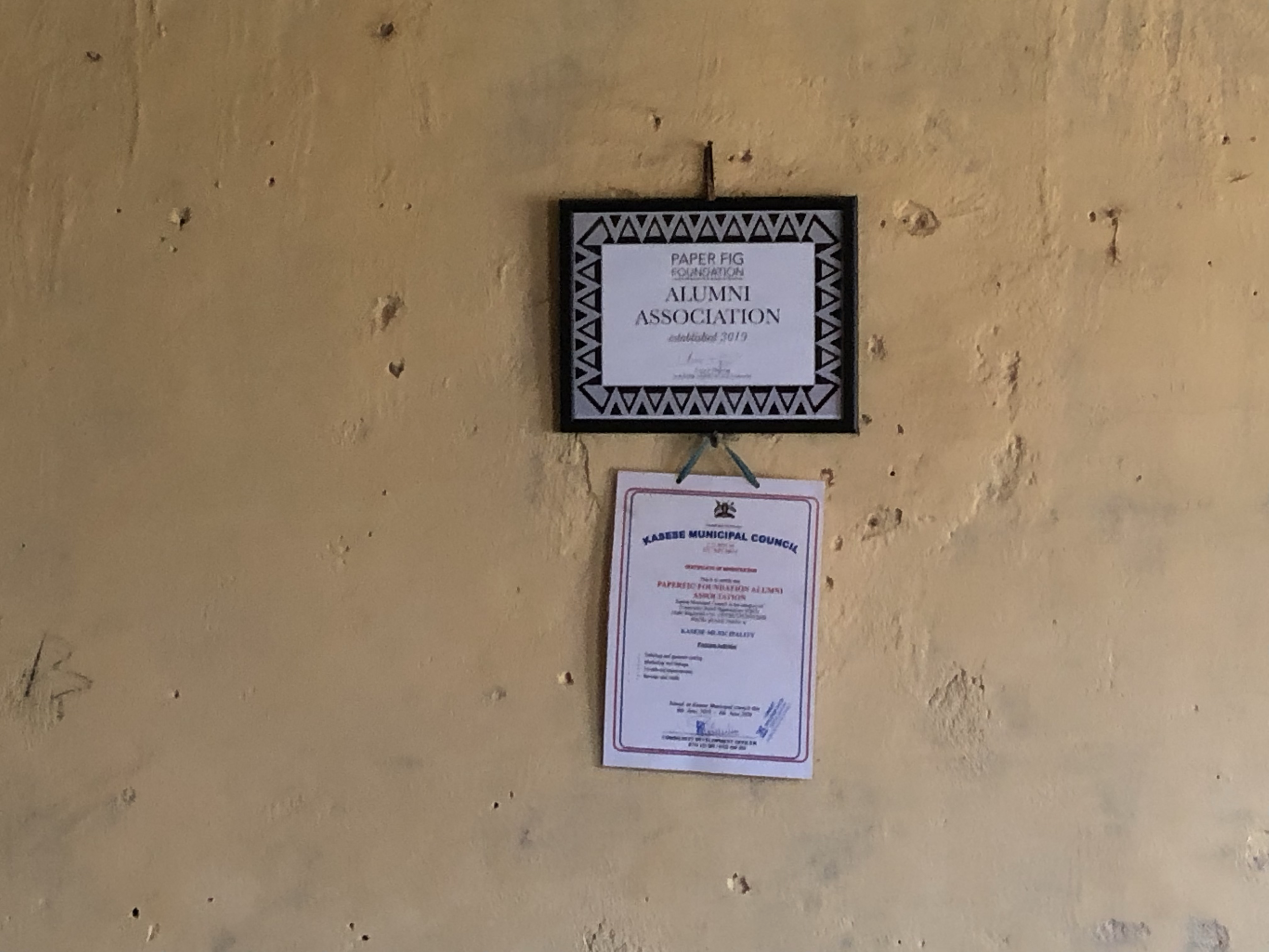 Wall with framed certificate and document, labeled "Paper Fig Foundation Alumni Association, established 2019" and "Kasese Municipal Council."
