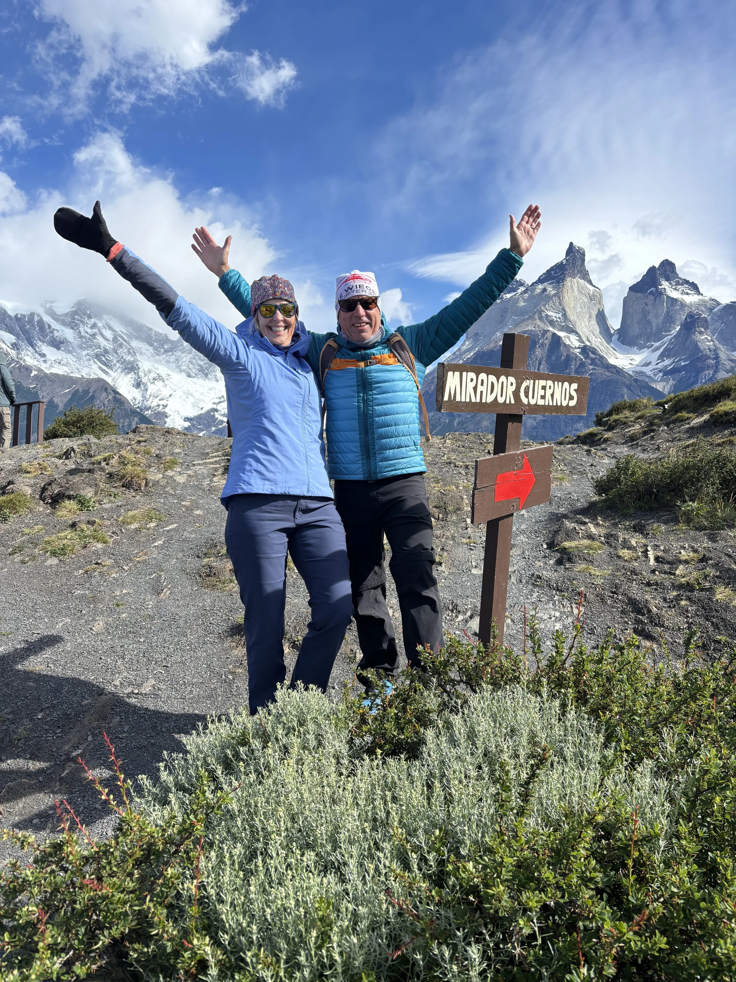 Two people in outdoor hiking gear celebrating at the Cuernos viewpoint in Torres del Paine National Park, with snow-capped mountains and a blue sky in the background.