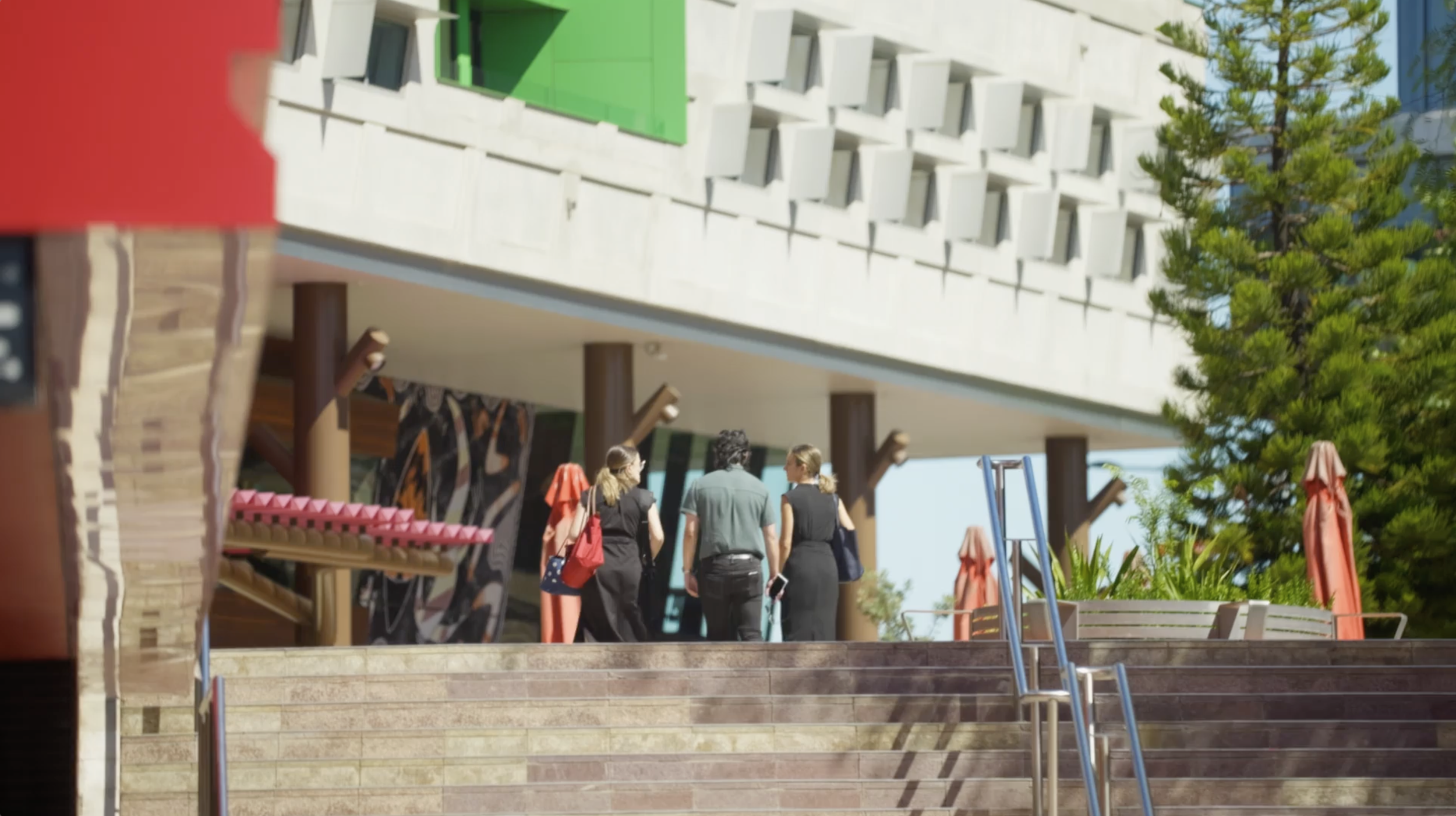 Three women and one man walking up the stairs outside a modern building with trees and umbrellas nearby.