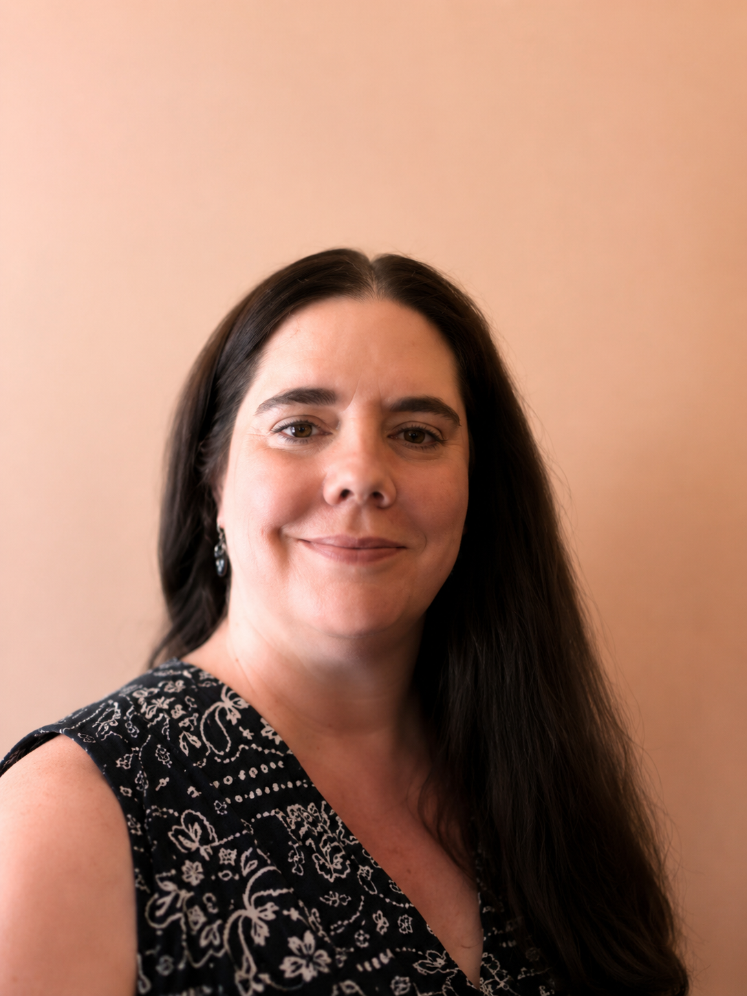 Close-up portrait of a woman with long dark hair, wearing earrings and a black dress with white floral patterns, smiling softly against a beige background.
