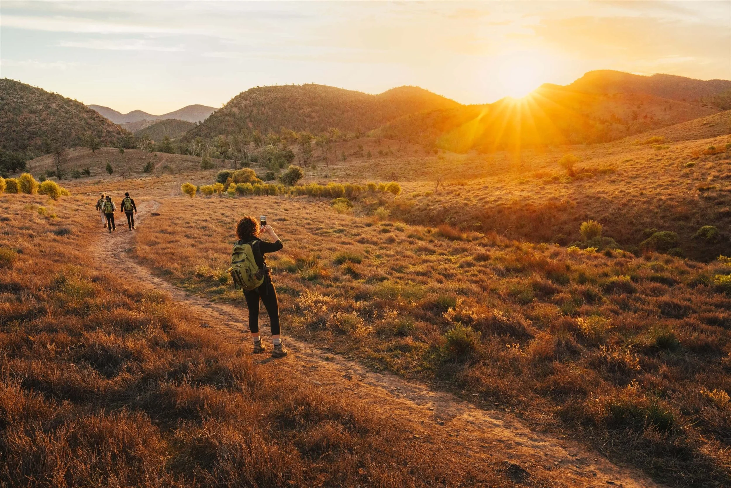 Group of hikers walking along a dirt trail in a hilly landscape at sunset, with one person taking a photo of the scenic view.