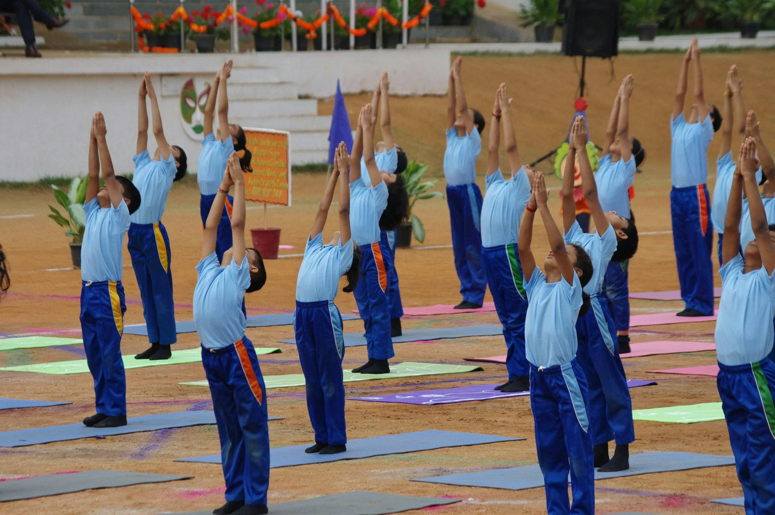 Group of school children practicing yoga outdoors on a sandy ground, wearing blue uniforms, with some standing on yoga mats, practicing yoga poses with raised hands. Decorations and a signboard are visible in the background.