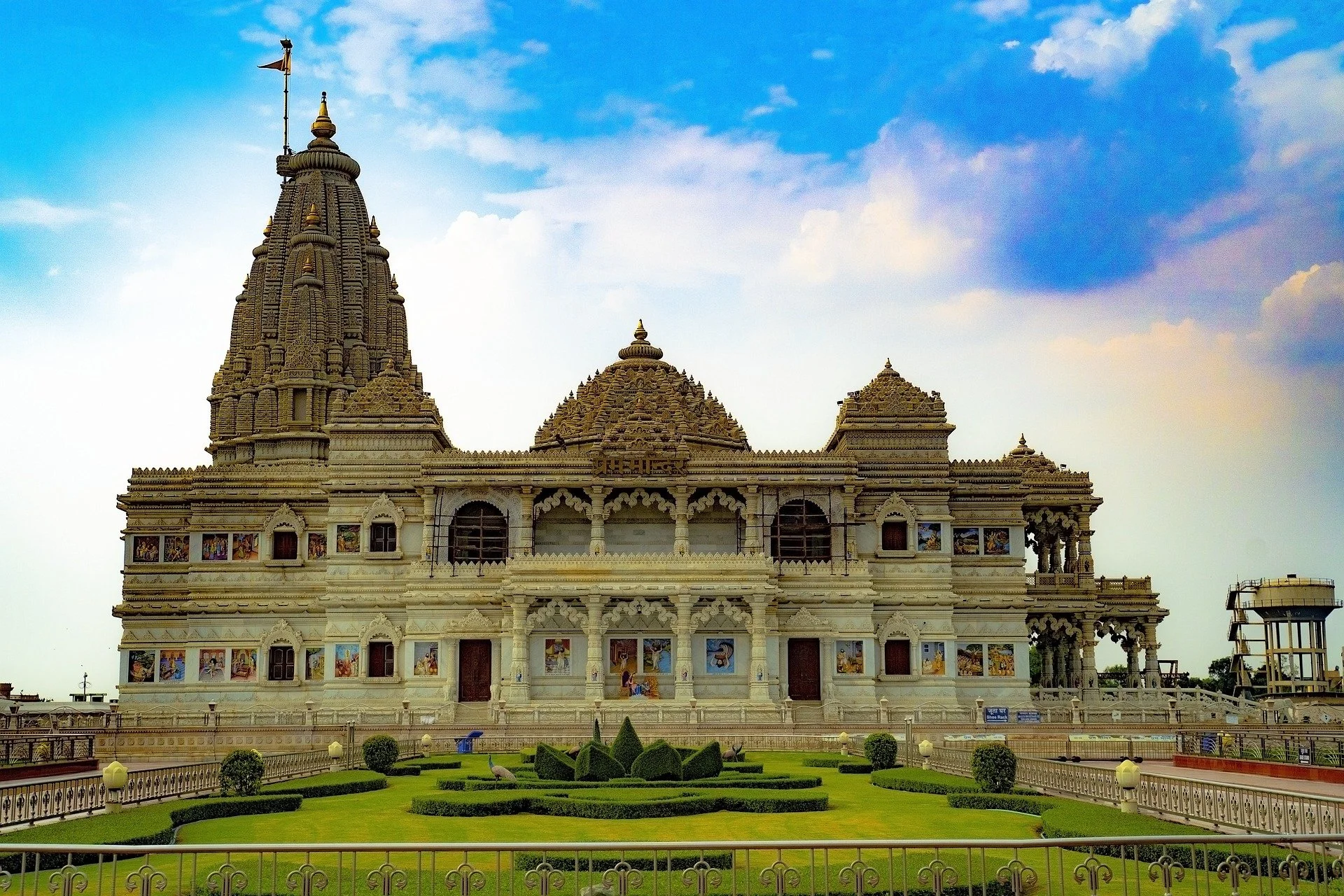 The image shows a large, ornate Hindu temple with intricate carvings, multiple domes, and a tall central spire topped with a flag. The temple is surrounded by well-maintained green lawns and a decorative fence, with the sky and clouds in the background.