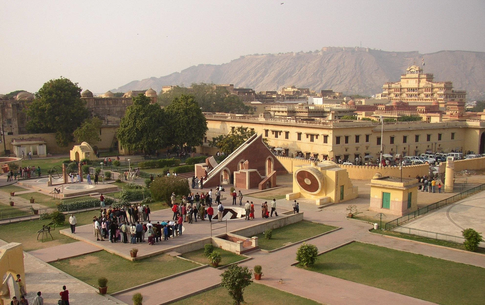 View of a historic monument with tourists and visitors, featuring architectural structures, green trees, and a mountain in the background.