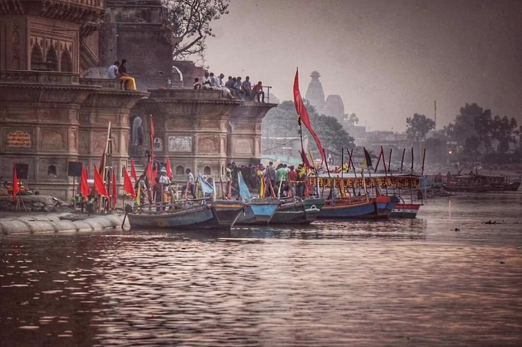 Boats docked along the riverbank near an old, ornate building with people gathered on the steps and on the nearby terrace, in a dusty, twilight setting.
