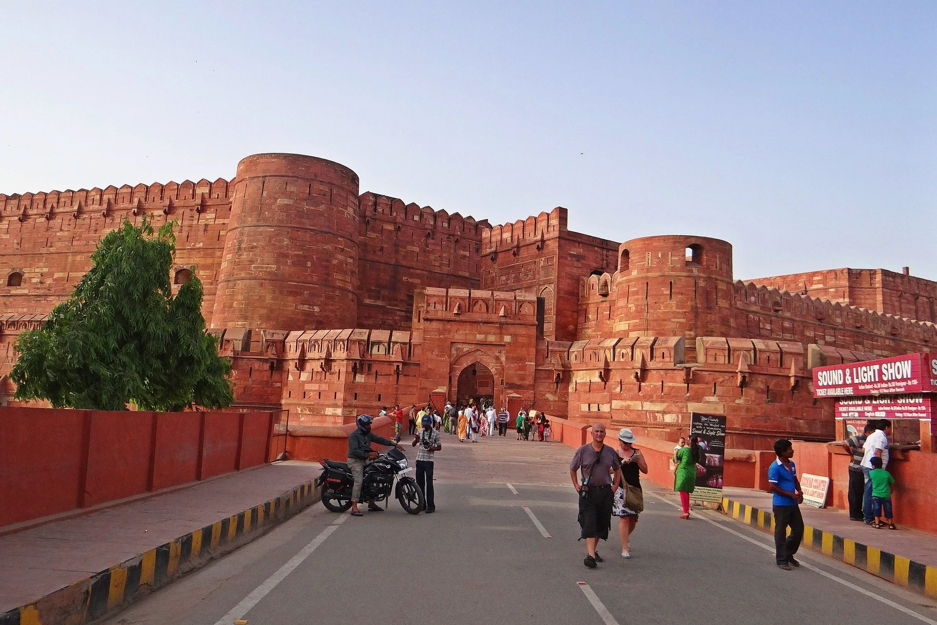 Red sandstone fortress with visitors walking near entrance, advertising sound and light show, and green tree in foreground.