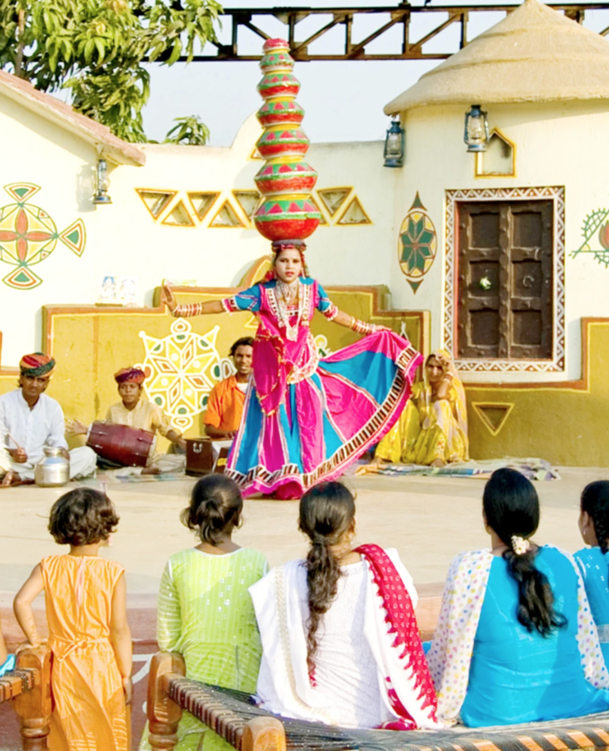 A dancer in colorful traditional Indian attire performing in front of an audience in an outdoor setting, with musicians playing in the background.