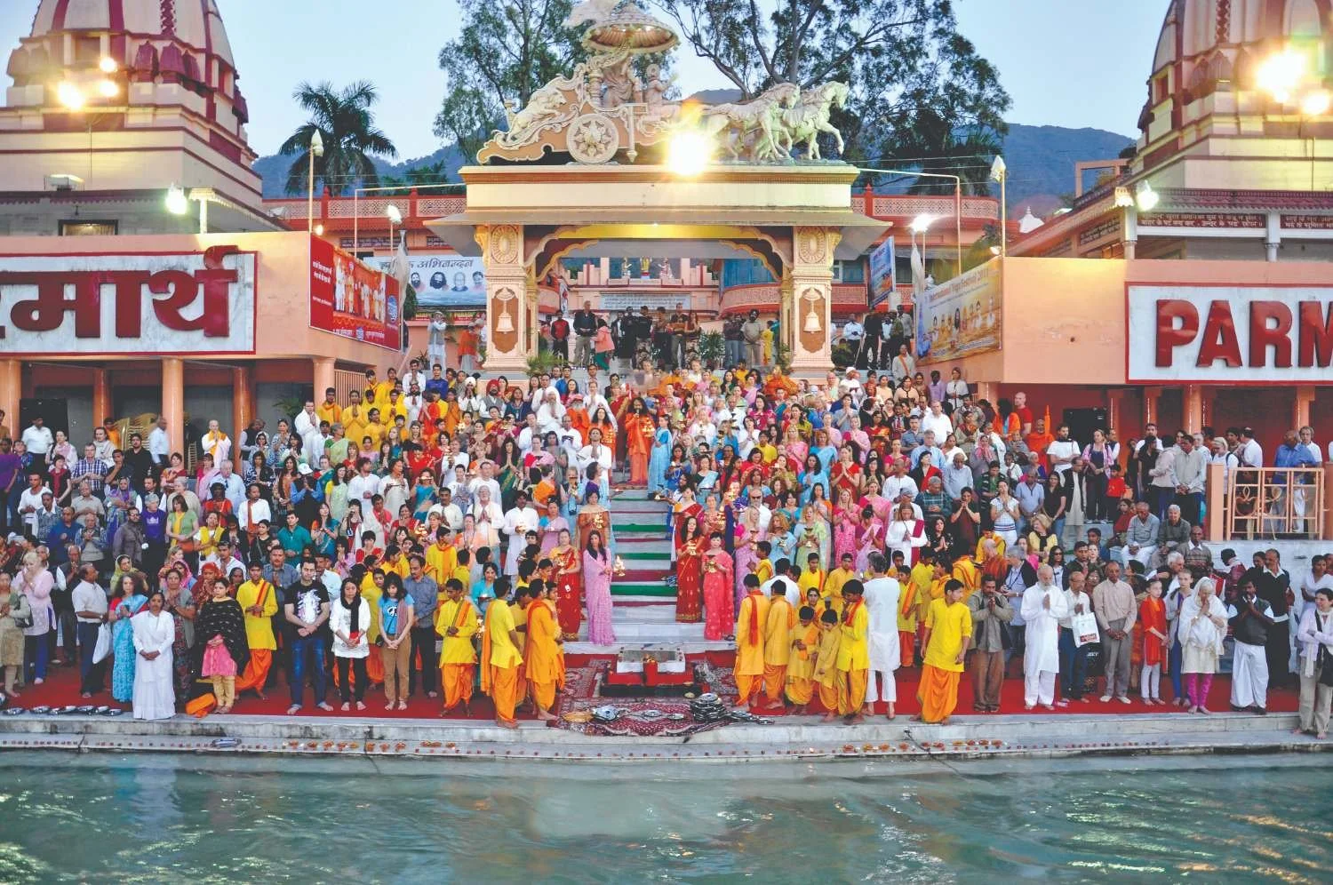 A large gathering of people at a Hindu temple near a water body, with many wearing colorful traditional clothing. The temple entrance features a gopuram with sculptures, and there are signs in Hindi and English. The scene appears to be during a festival or religious event.