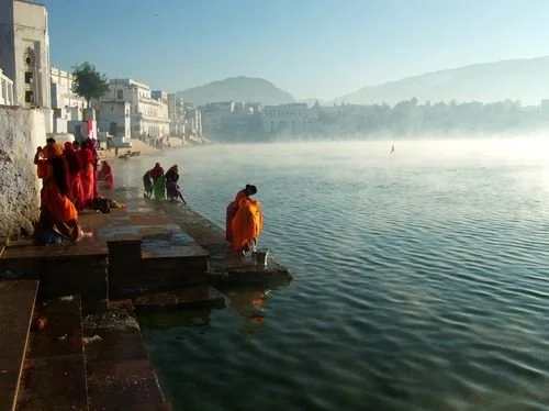 People dressed in colorful clothing gather by the riverbank with historic buildings lining the shore, mist rising over the water and mountains in the background.