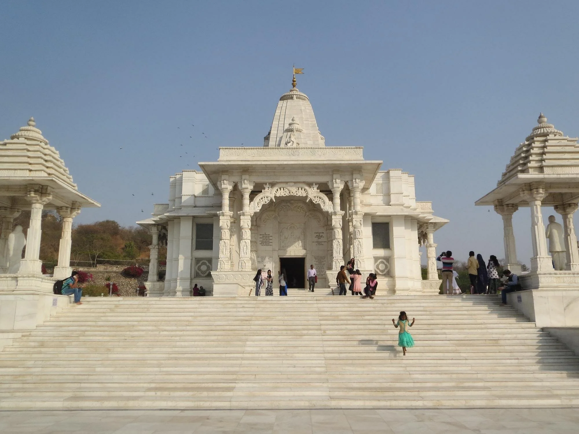 White marble Hindu temple with steps leading up to the entrance, surrounded by smaller structures with statues, and people walking and sitting around it, under a clear blue sky.