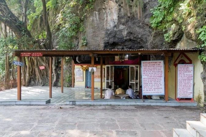 Small Hindu shrine with people praying inside, situated at the base of a rocky hillside with greenery, protected by a metal roof and open sides.