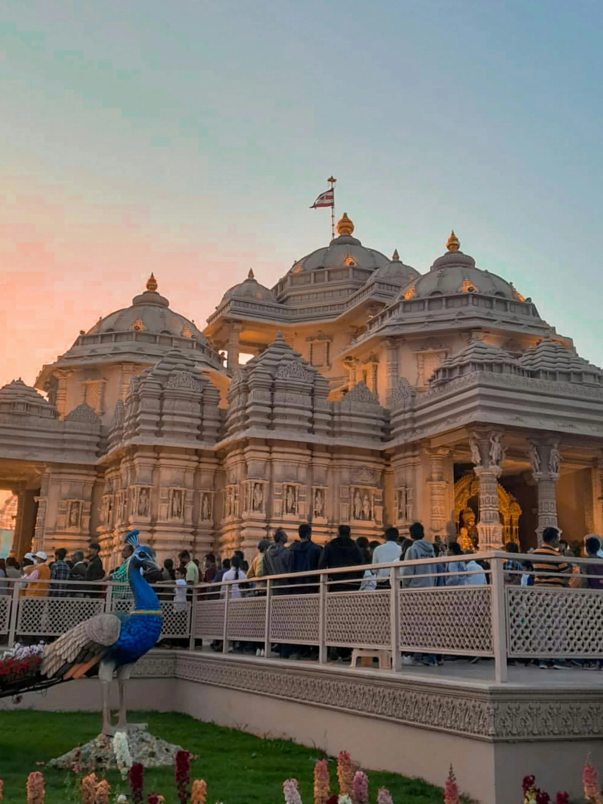 Hindu temple with multiple domes, people gathered around, and a large peacock statue in the foreground during sunset.