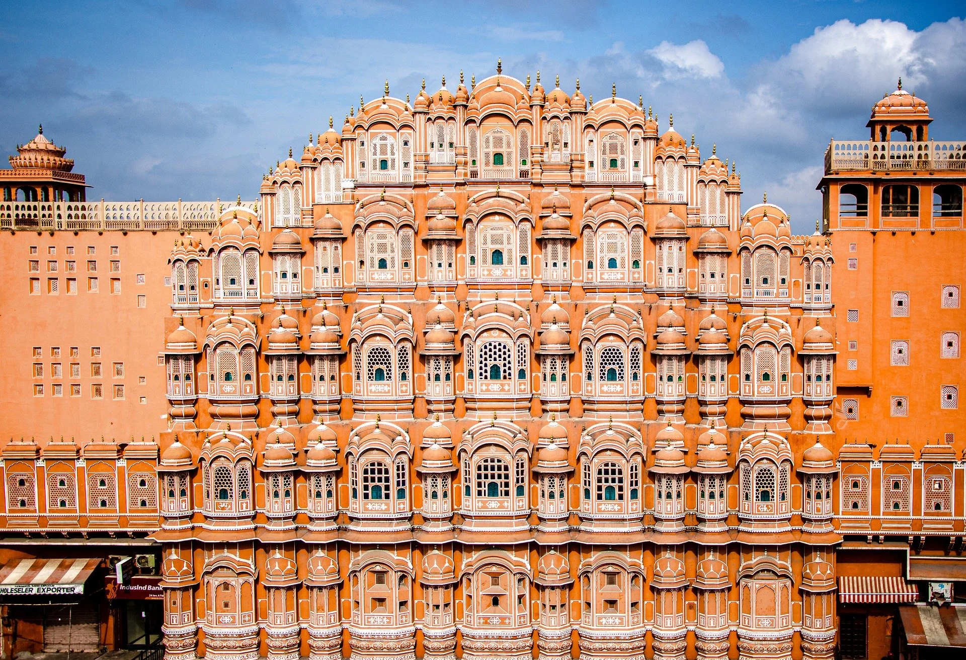Front view of Hawa Mahal, an ornate pink sandstone palace with many small windows and domed arches, under a partly cloudy blue sky.
