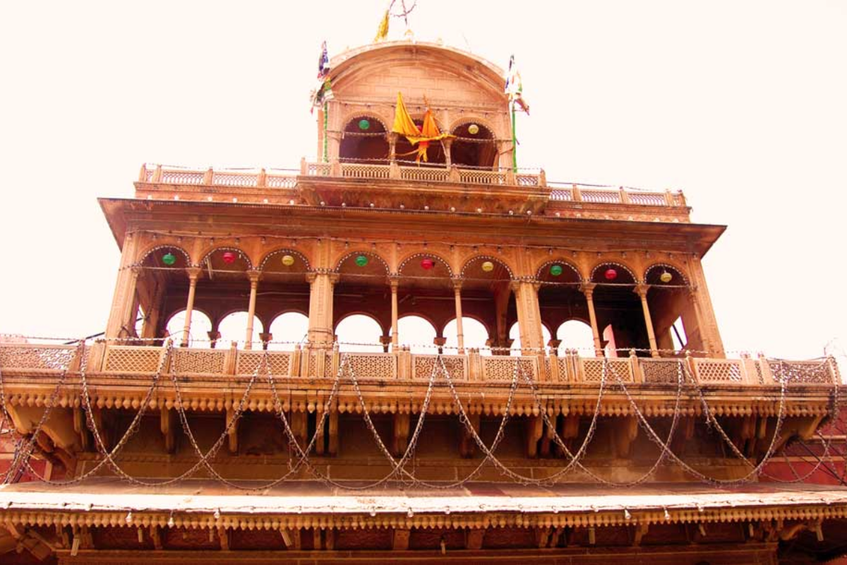 A multi-story Hindu temple made of reddish stone or brick, decorated with colorful flags and string lights, with multiple arches and balconies.
