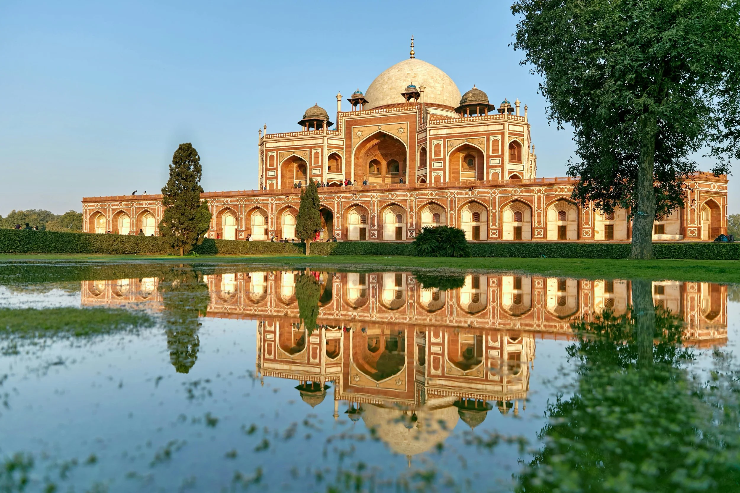The Taj Mahal with its reflection in a water pool in front, surrounded by green trees and a clear blue sky.