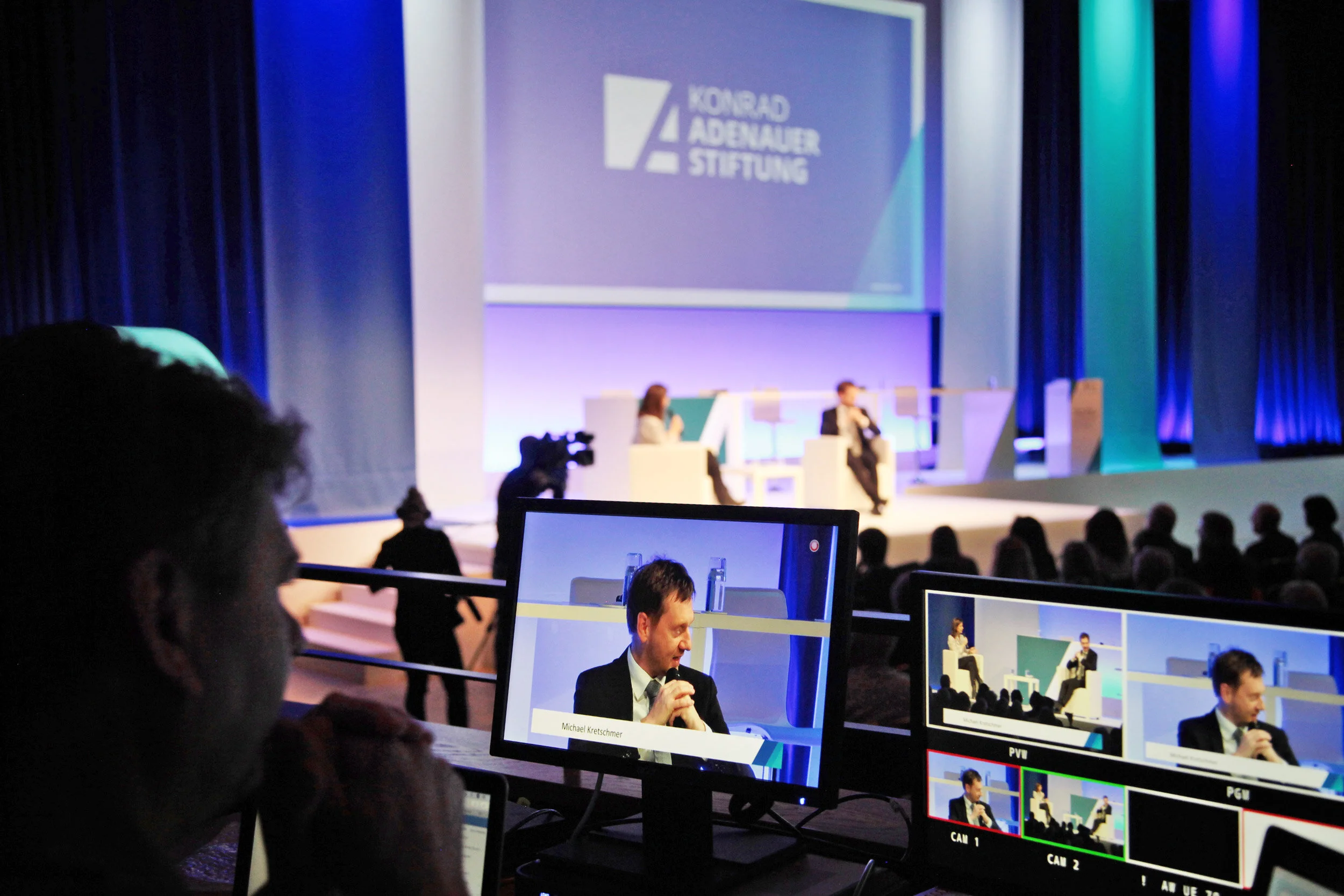 Foto von zwei Bildschirmen, die den Talk im Hintergrund zeigen, wo gerade Michael Kretschmer zu sehen ist, wie er das Mikrofon hält