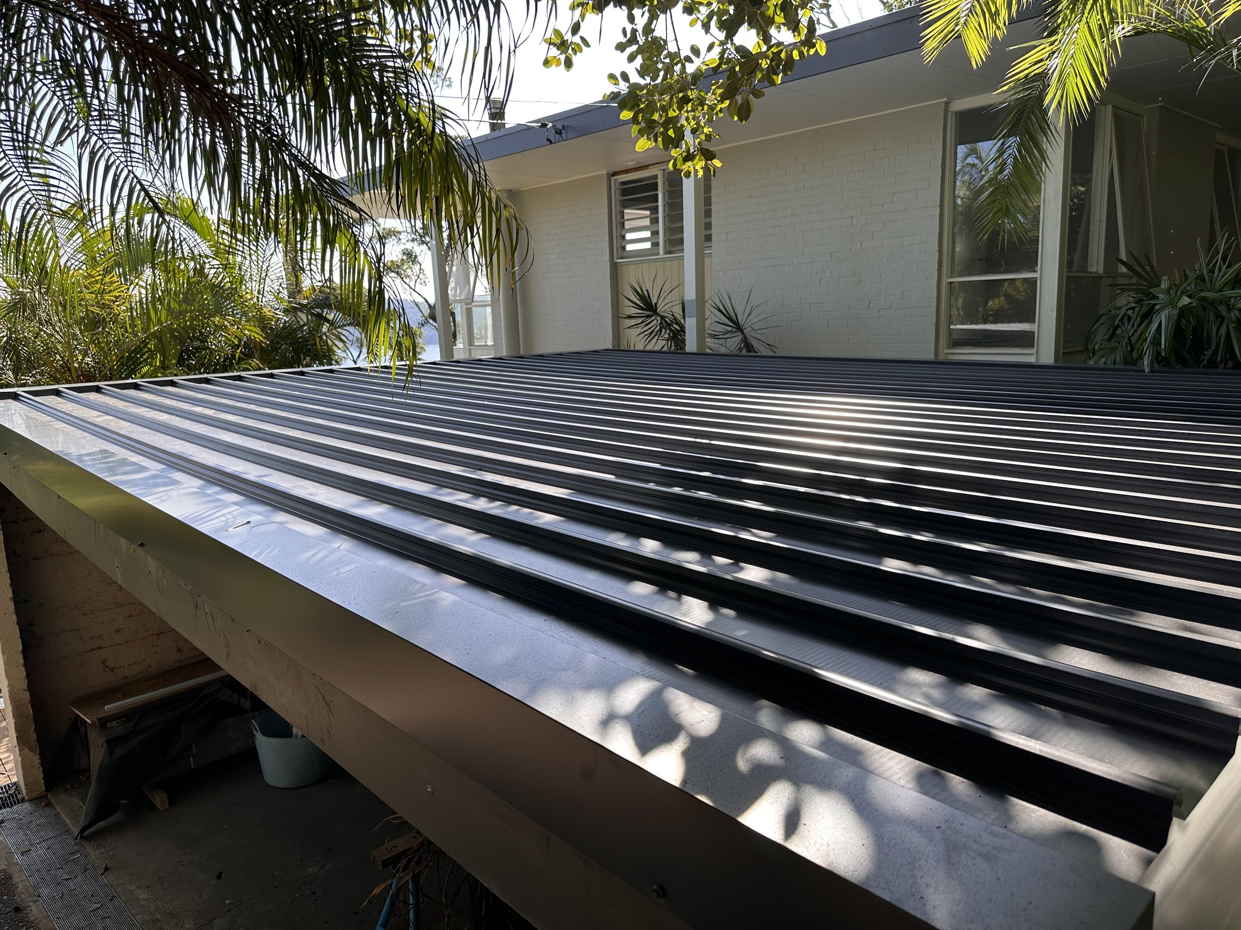 A metal roof with black standing seam panels on a residential building surrounded by palm trees and greenery.
