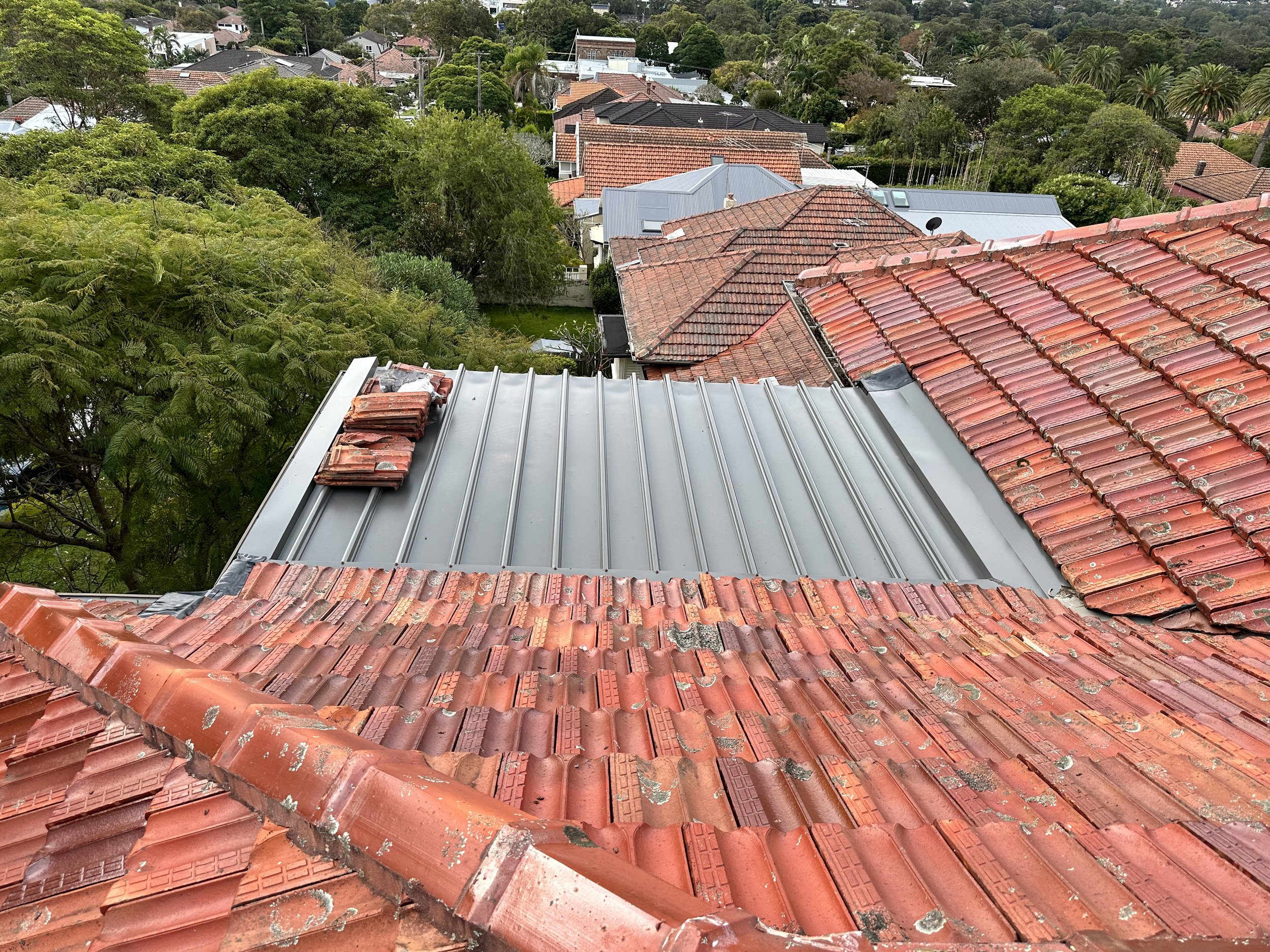 View of rooftops with red tiles and a section of new metal roofing on an elevated position over a neighborhood with trees and houses.