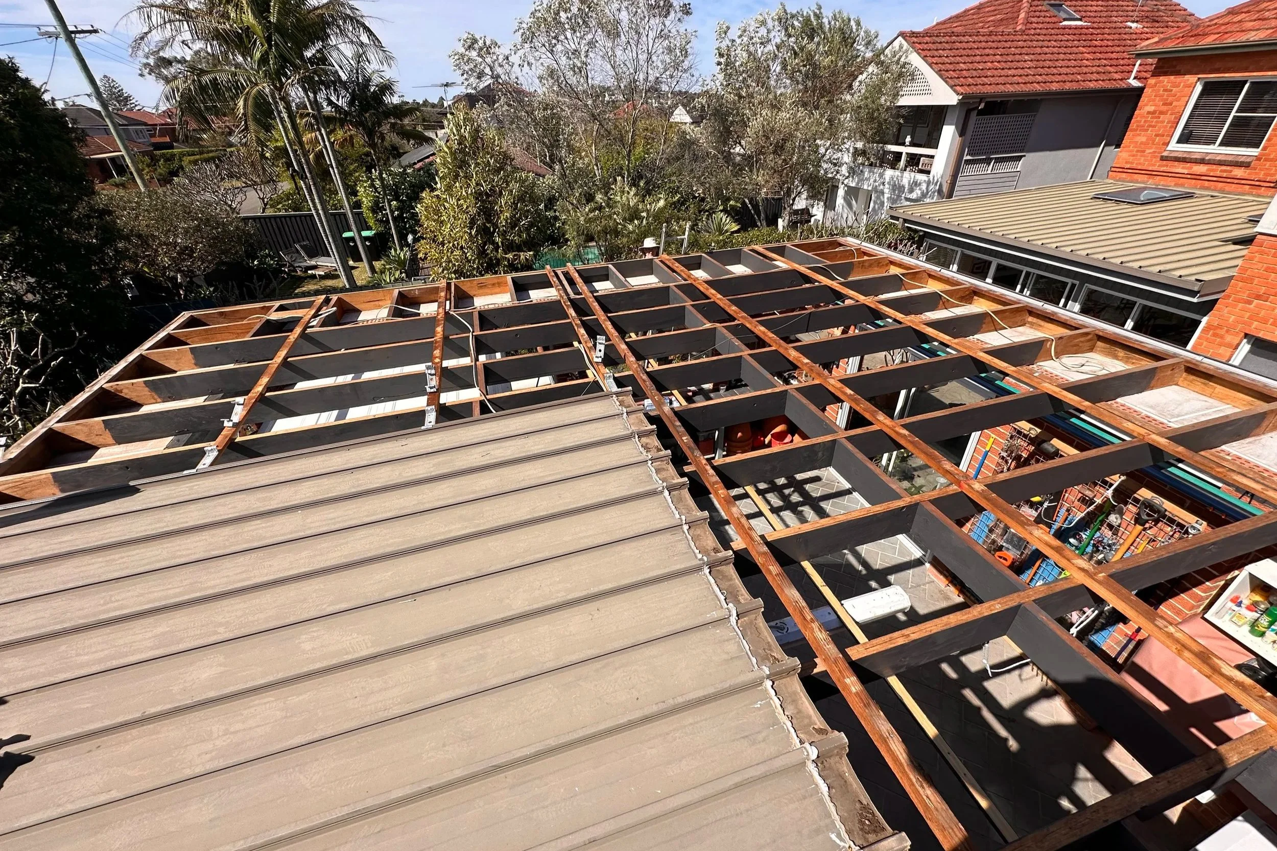 A rooftop under construction with wooden framing and black roofing panels. The backdrop shows neighboring houses with various roof styles, trees, and utility poles.