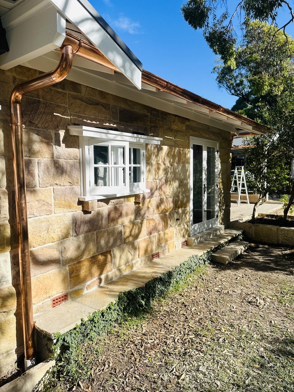 Stone house exterior with white window and glass door, copper downspouts, small garden bed, trees, clear blue sky, and ladder in the background.