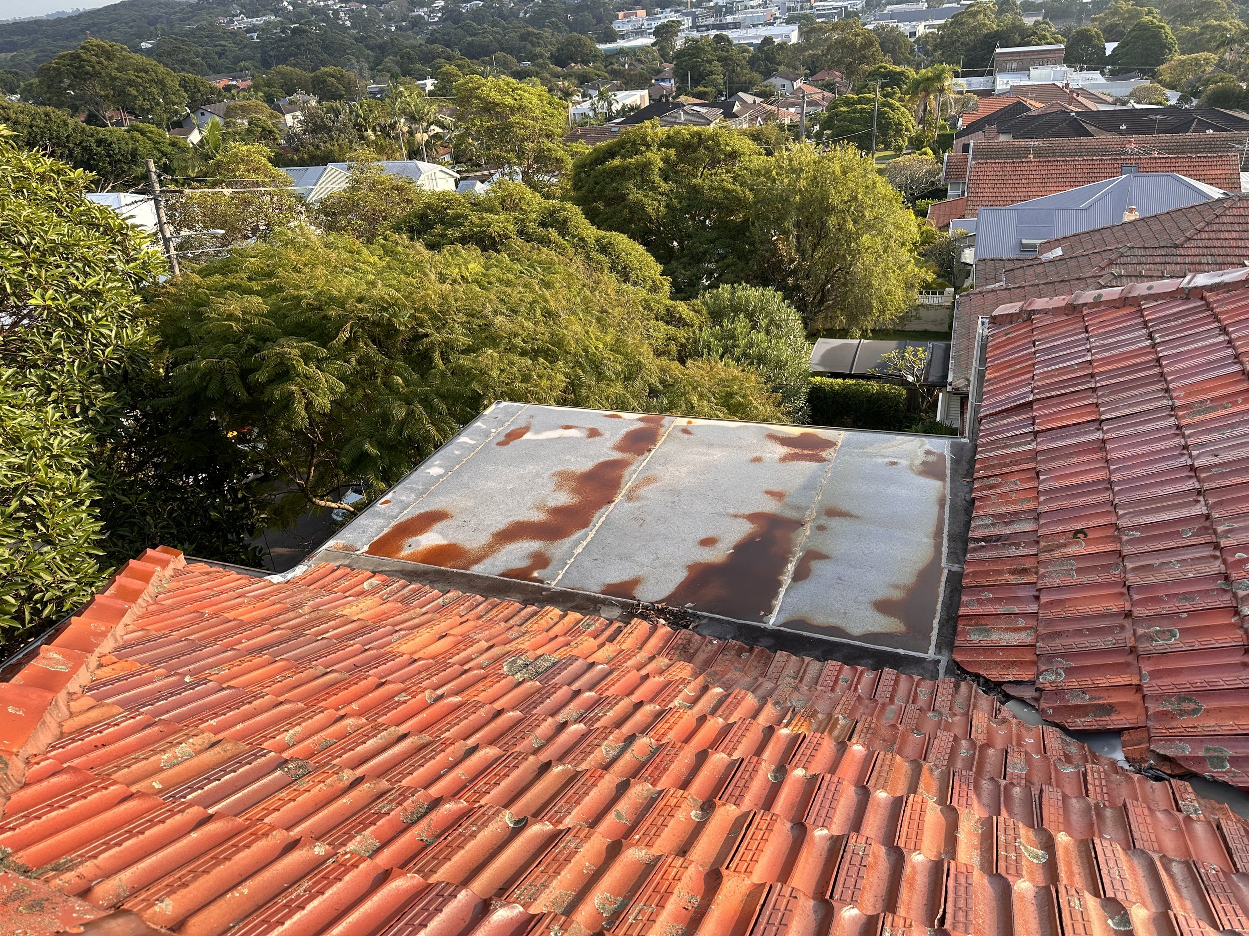 View of a rooftop with red tiles, next to a flat metal roof with rust patches, overlooking a neighborhood with trees and houses.