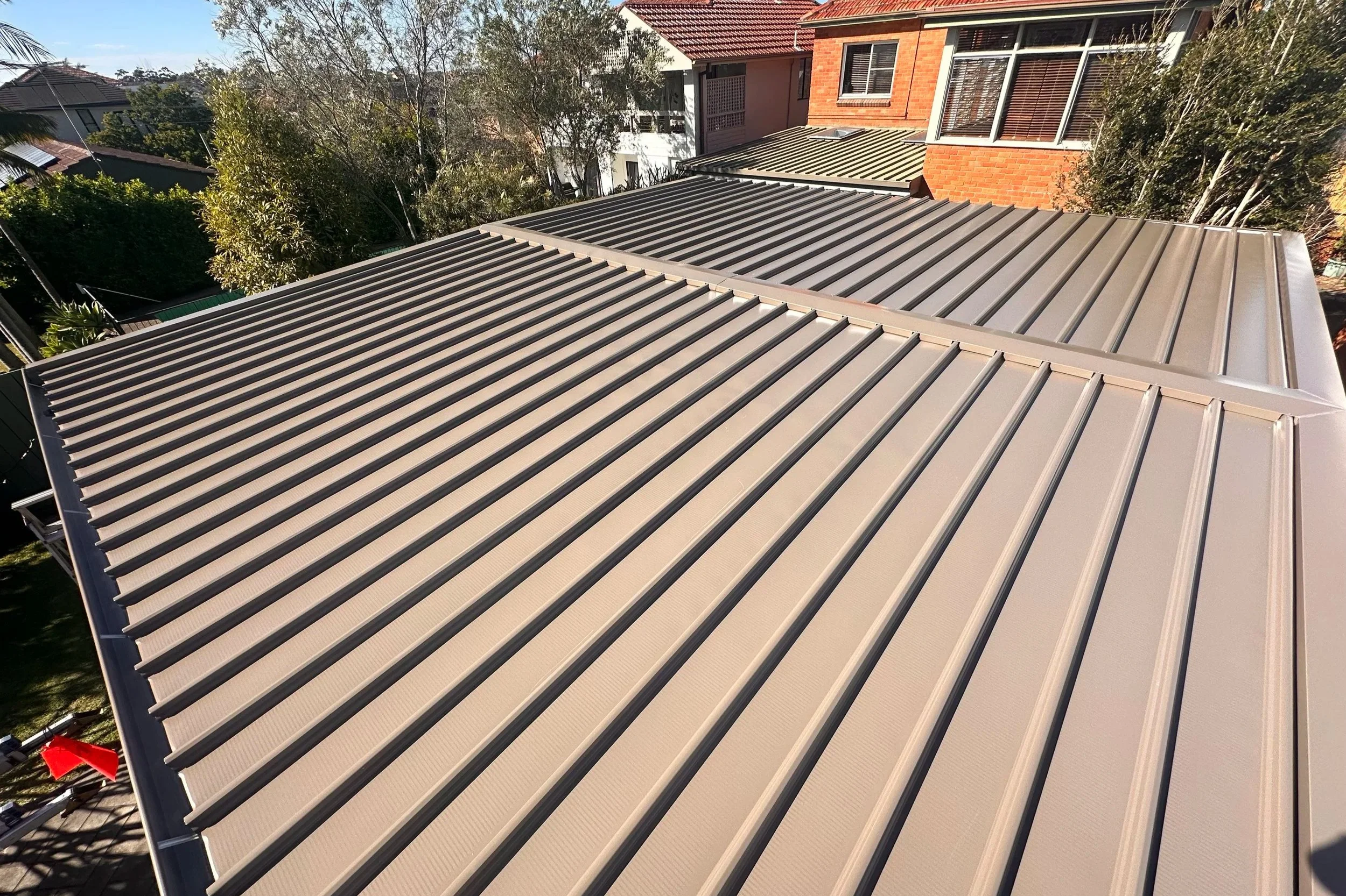 Aerial view of a house roof with metal roofing panels, some of which are beige and others gray, with neighboring houses and trees in the background.