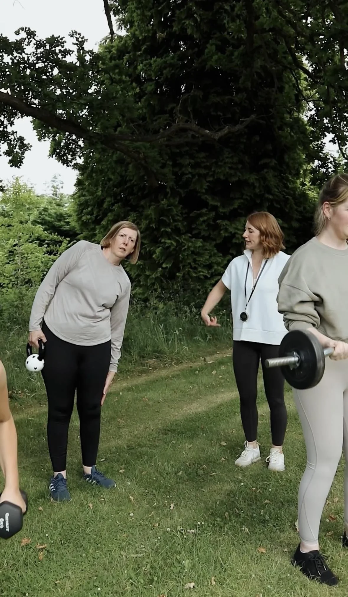 Three women outdoors near a large tree, one holding a small weight, another with a stopwatch around her neck, the third lifting a dumbbell.