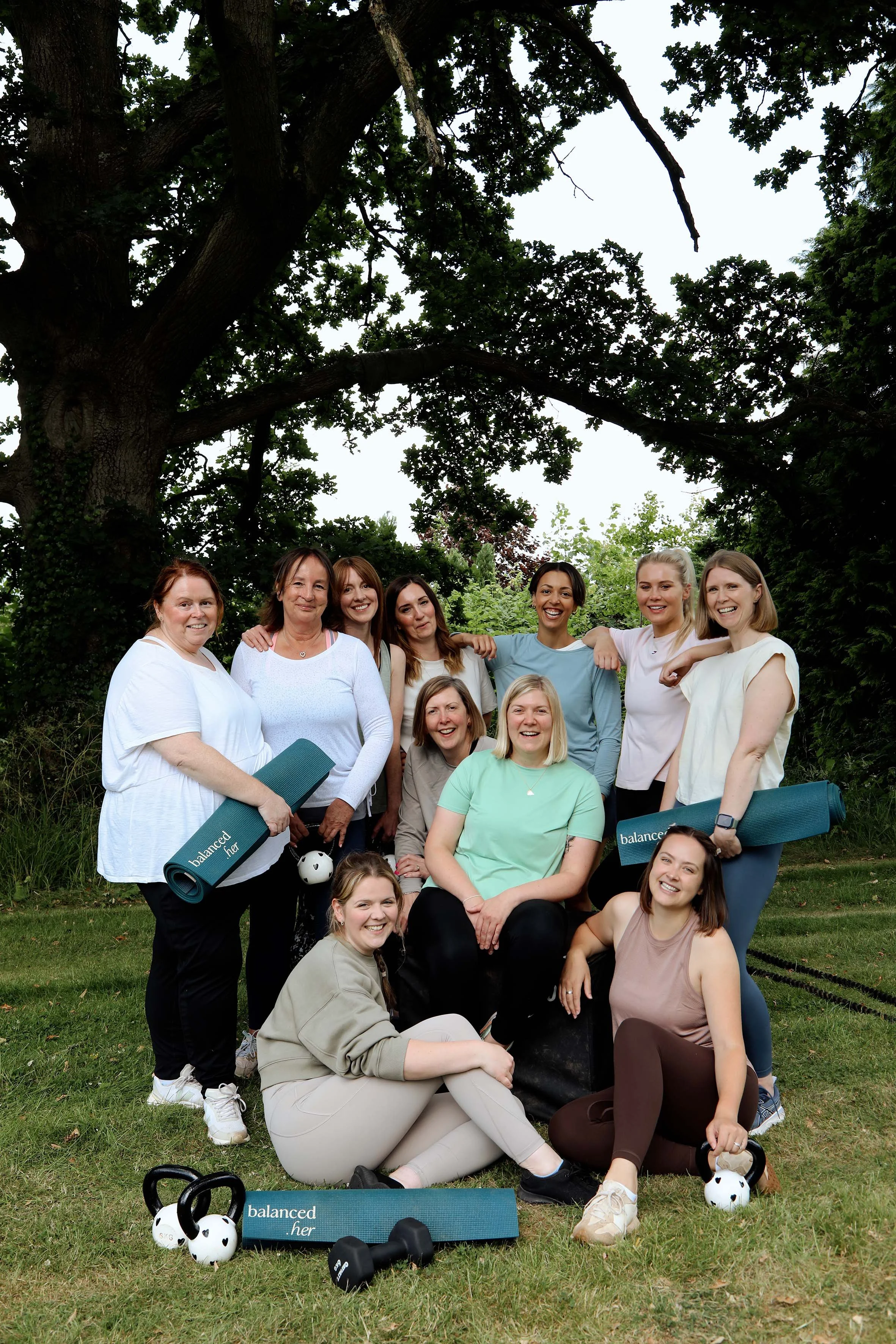 Group of smiling women and young women standing and sitting outdoors on grass in front of a large tree, some holding yoga mats and fitness equipment including kettlebells and a medicine ball, during daytime.