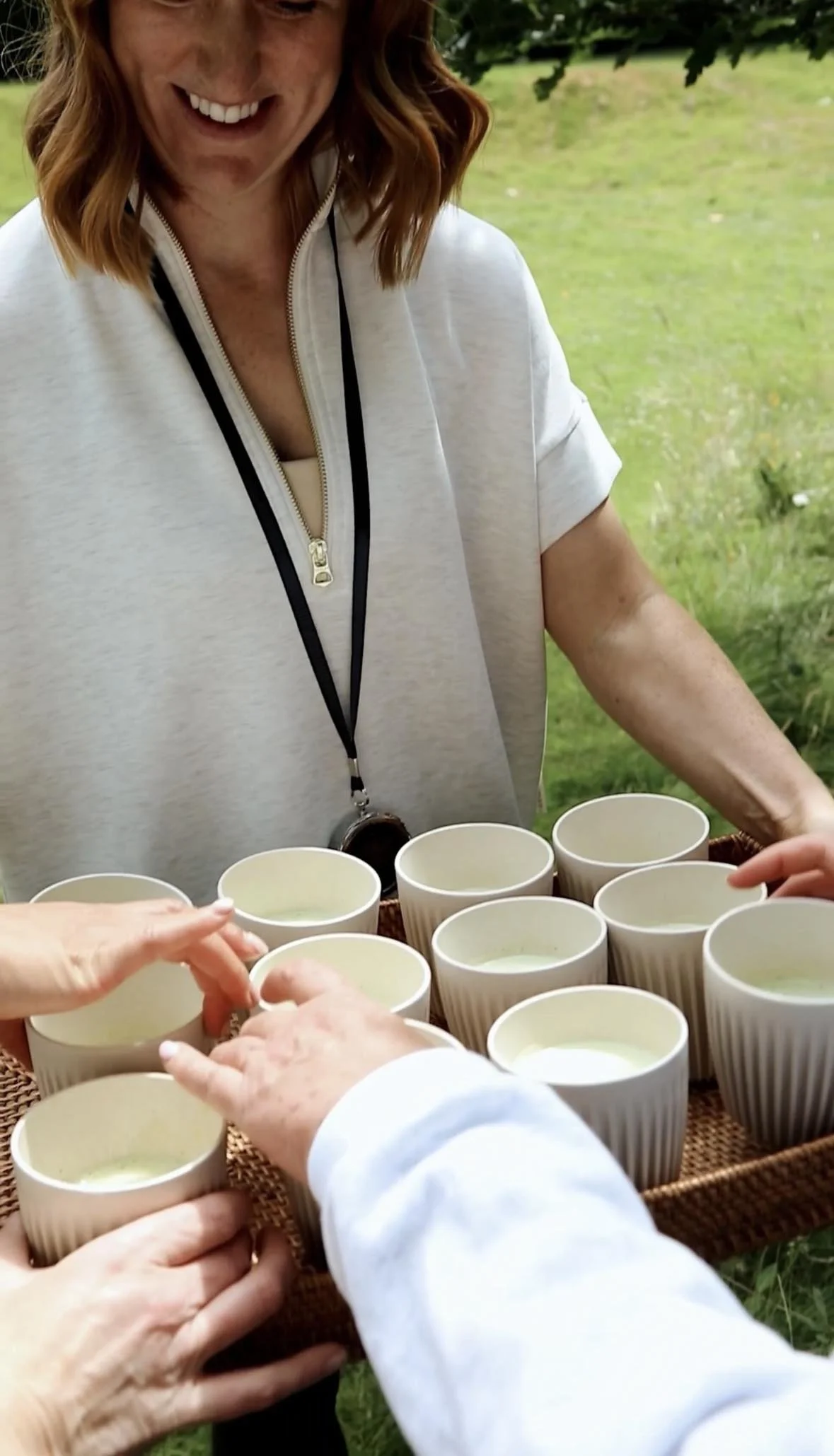 A woman with red hair, wearing a white zip-up top and a lanyard, is serving or handing out white cups filled with a light-colored beverage, with people reaching for the cups. They are outdoors, with greenery and trees in the background.