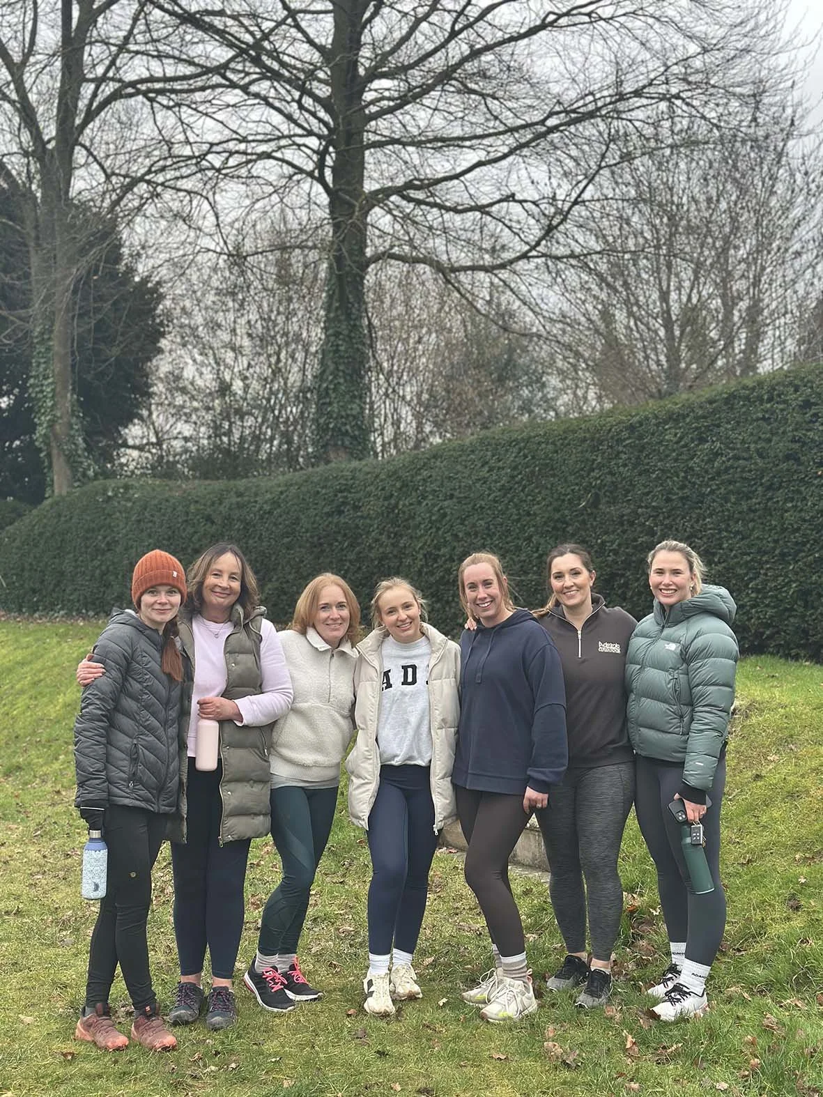 A group of seven women standing outdoors in front of a hedge and trees on a cloudy day, dressed in athletic and casual clothing, smiling for the photo.