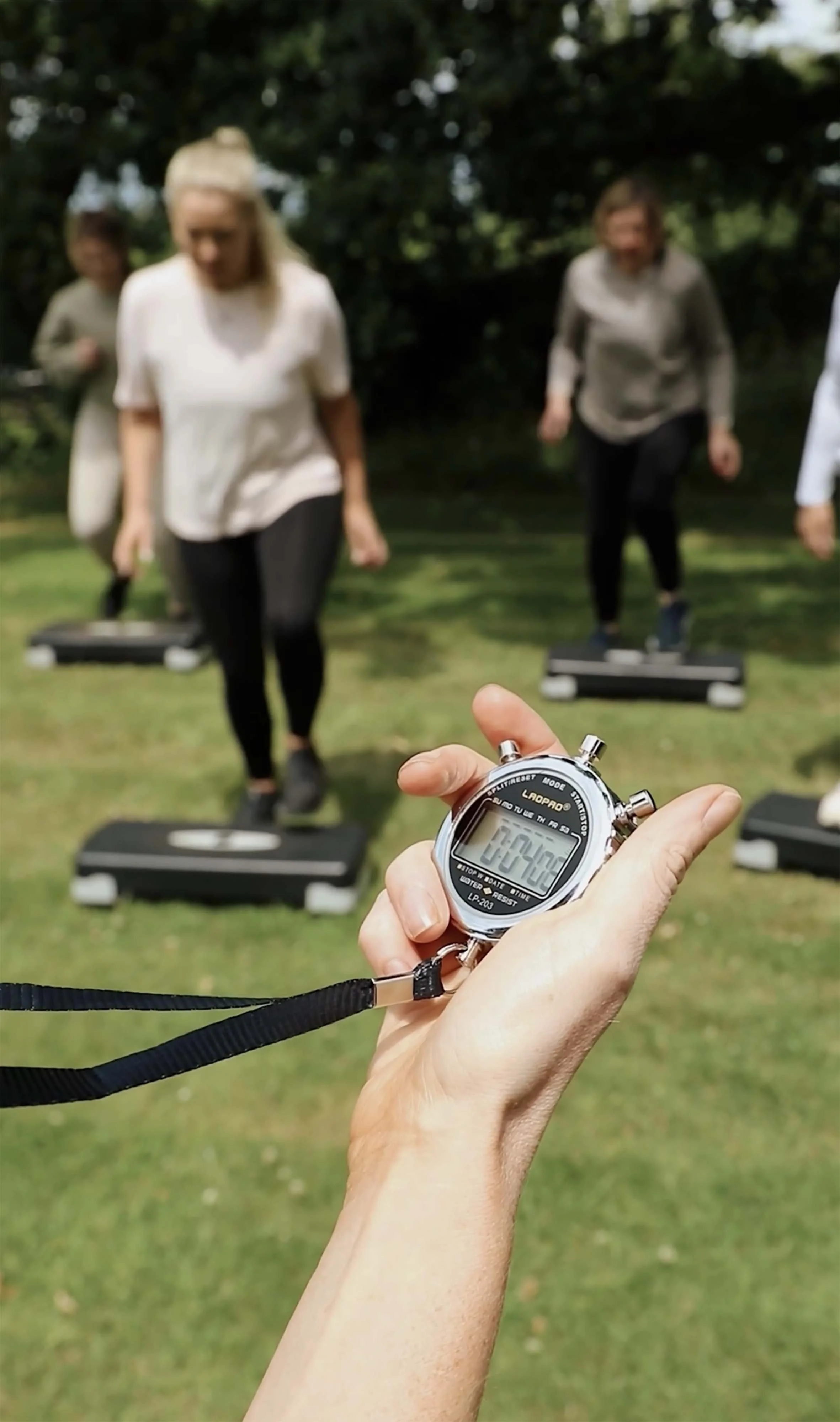 A hand holding a stopwatch in the foreground, with three women using step platforms outdoors in the background.