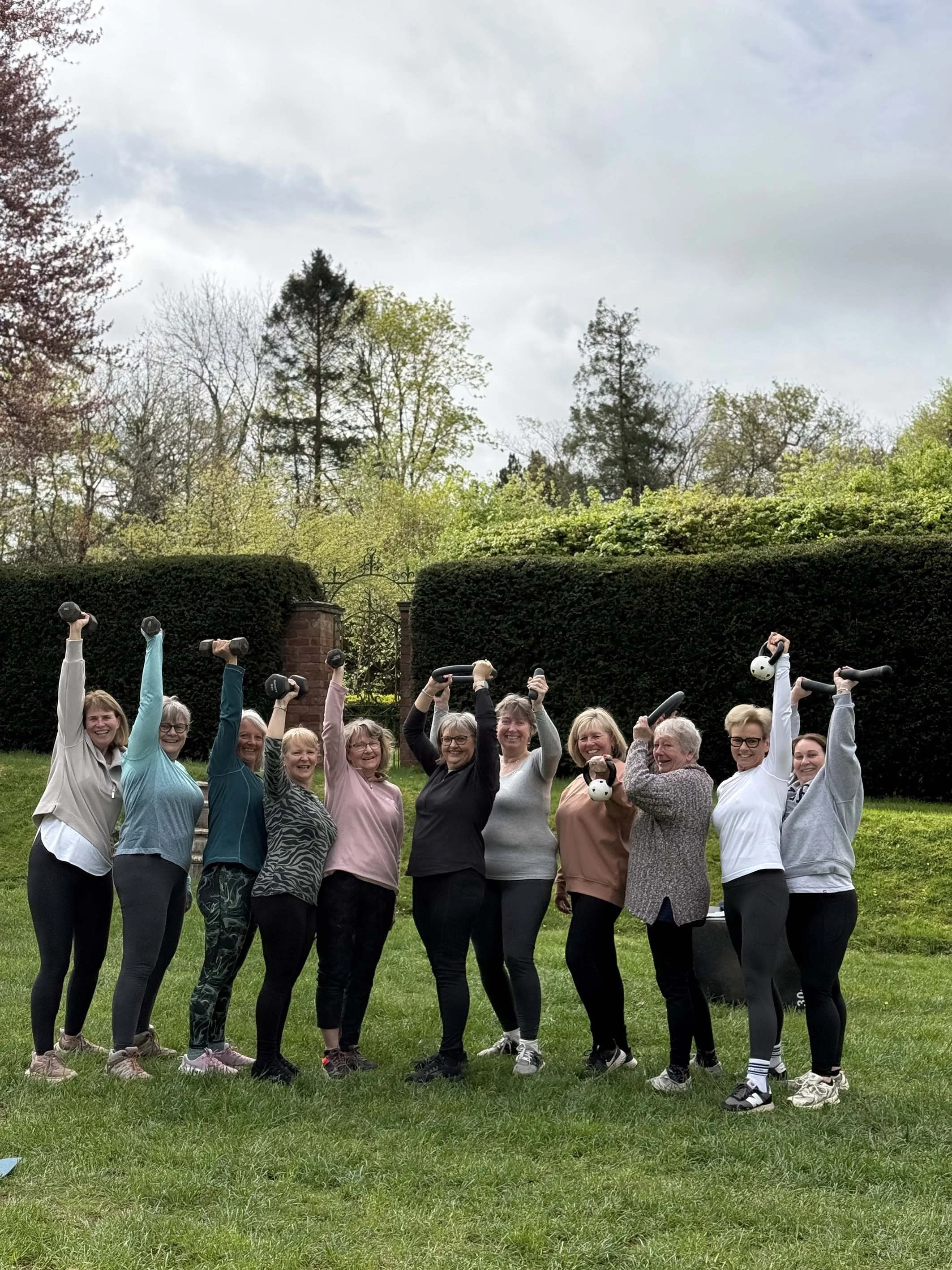 A group of women standing outdoors on grass, smiling and holding dumbbells and kettlebells. They are in front of a green hedge with trees and a cloudy sky behind them.