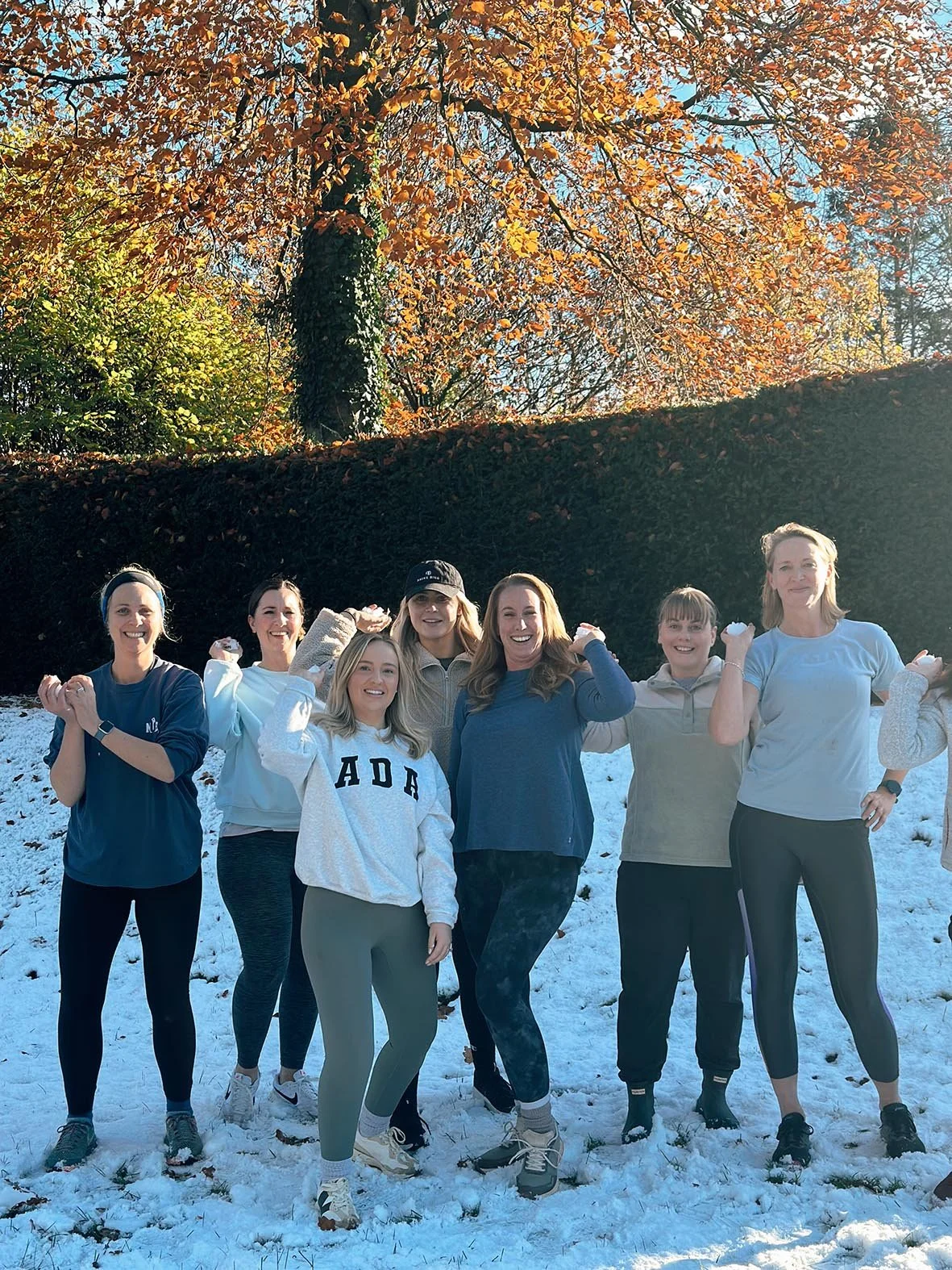 A group of seven women standing outside on snow-covered ground, raising their arms in a fitness pose, with colorful autumn trees in the background.