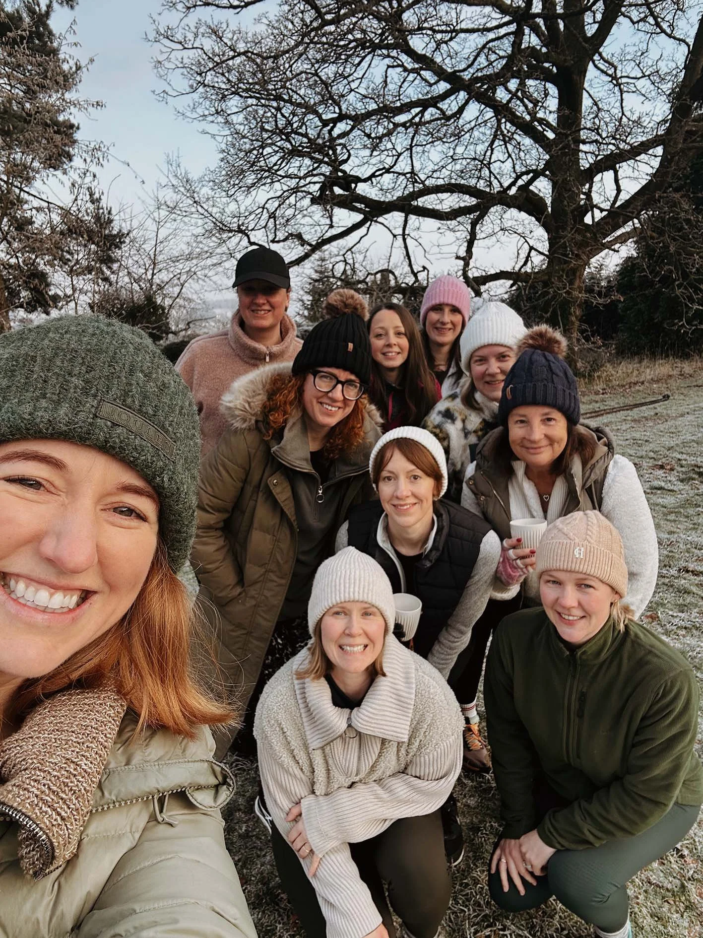 A group of women outdoors in a chilly environment, all dressed in warm clothing, smiling for a selfie with a large tree and winter landscape in the background.