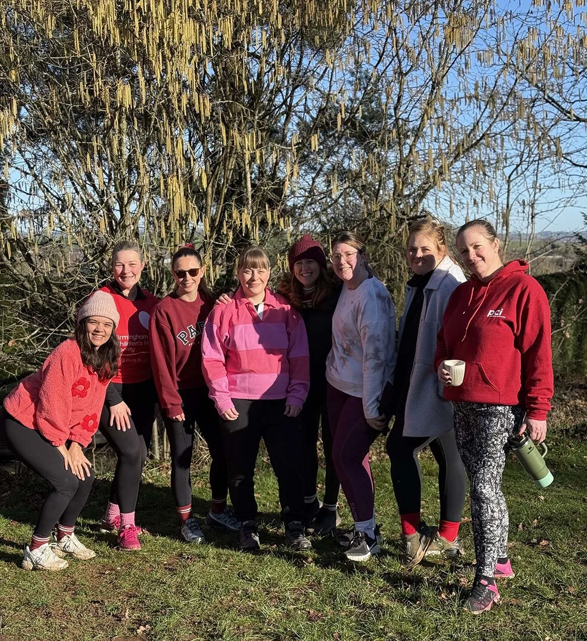 Group of eight women standing outdoors on a sunny day, smiling in front of a large tree with hanging catkins, wearing casual athletic clothing in pink, red, and gray, with some holding water bottles and a mug.