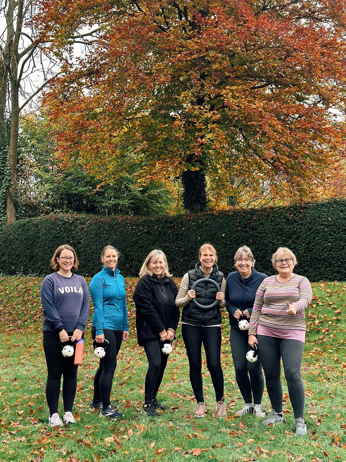 Six women dressed in athletic clothing standing outdoors on a grassy area with fallen autumn leaves, holding kettlebells, and smiling at the camera, with large trees and a trimmed hedge in the background.