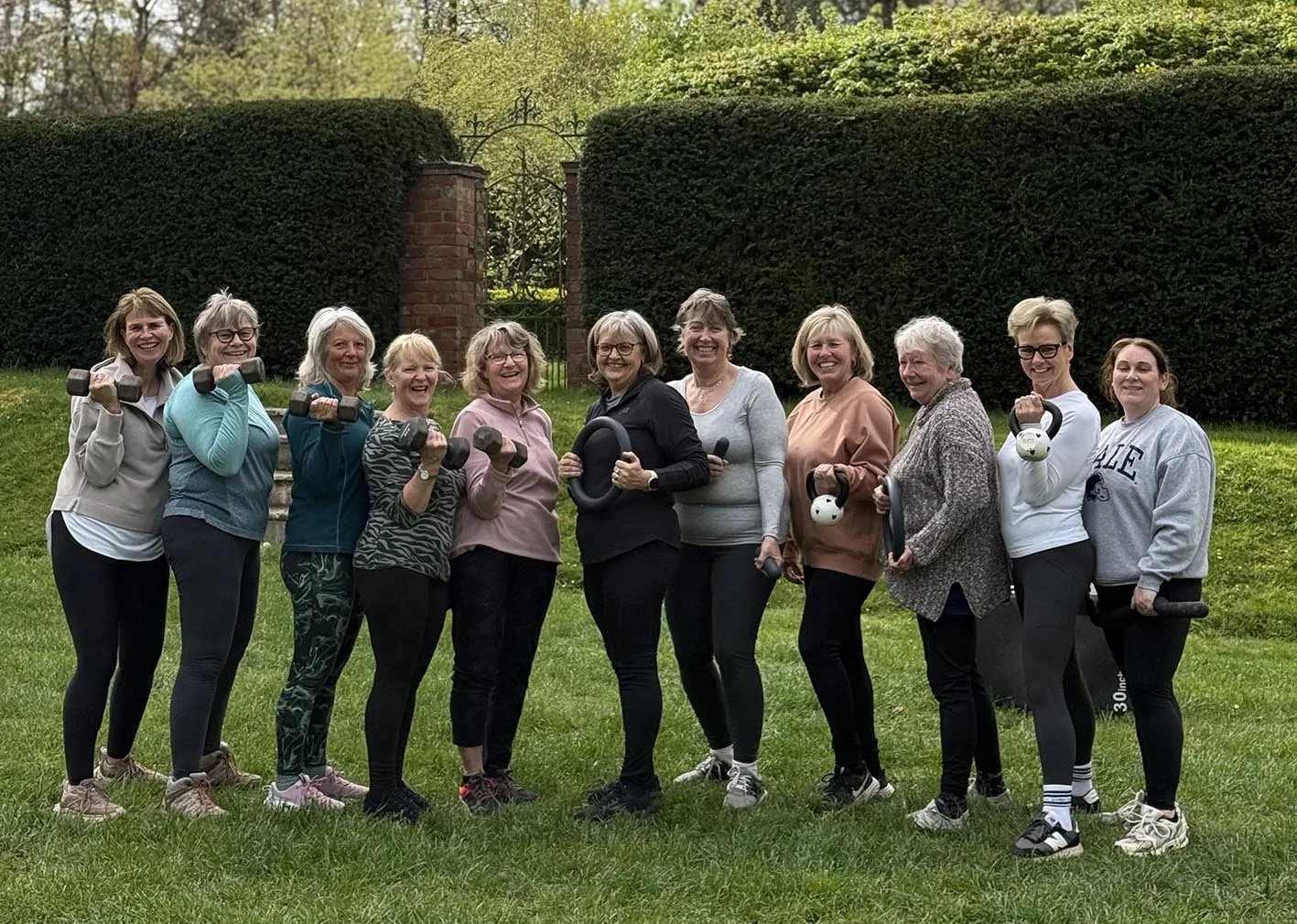 Group of women doing a workout with dumbbells and kettlebells outdoors on a grassy lawn with green bushes and trees in the background.
